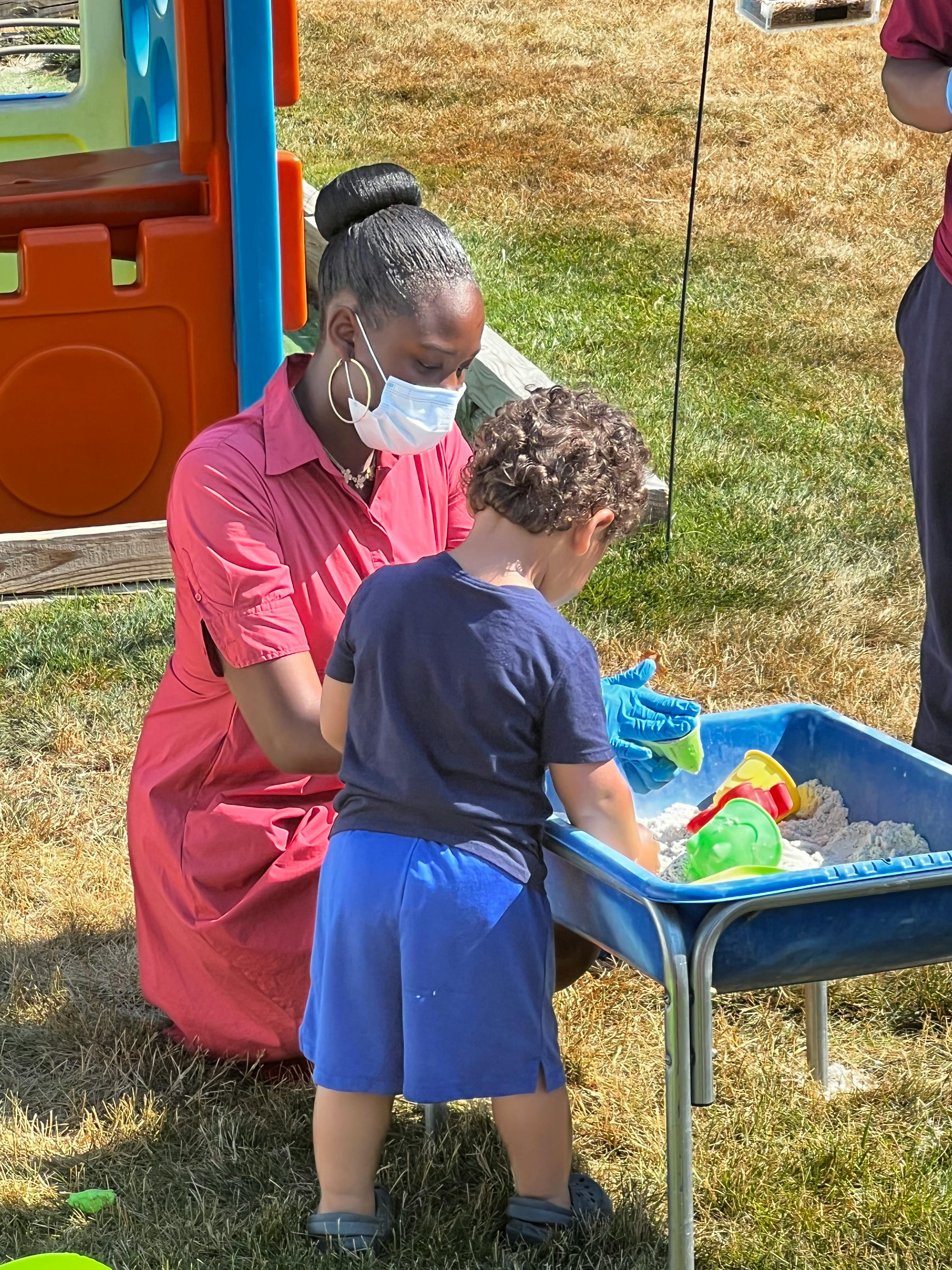 Person in mask kneeling next to child playing in sand table