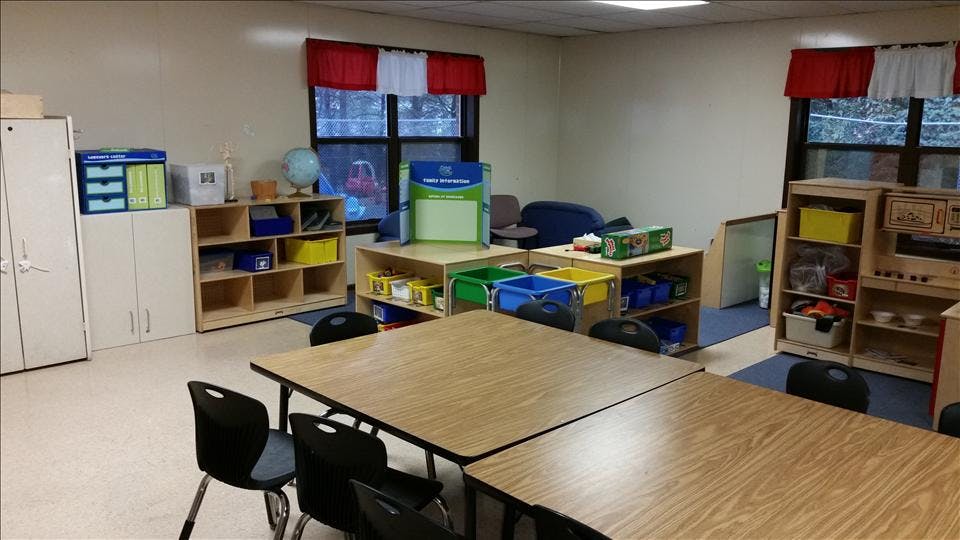 An empty classroom with small tables and chairs, shelves, and red and white curtains