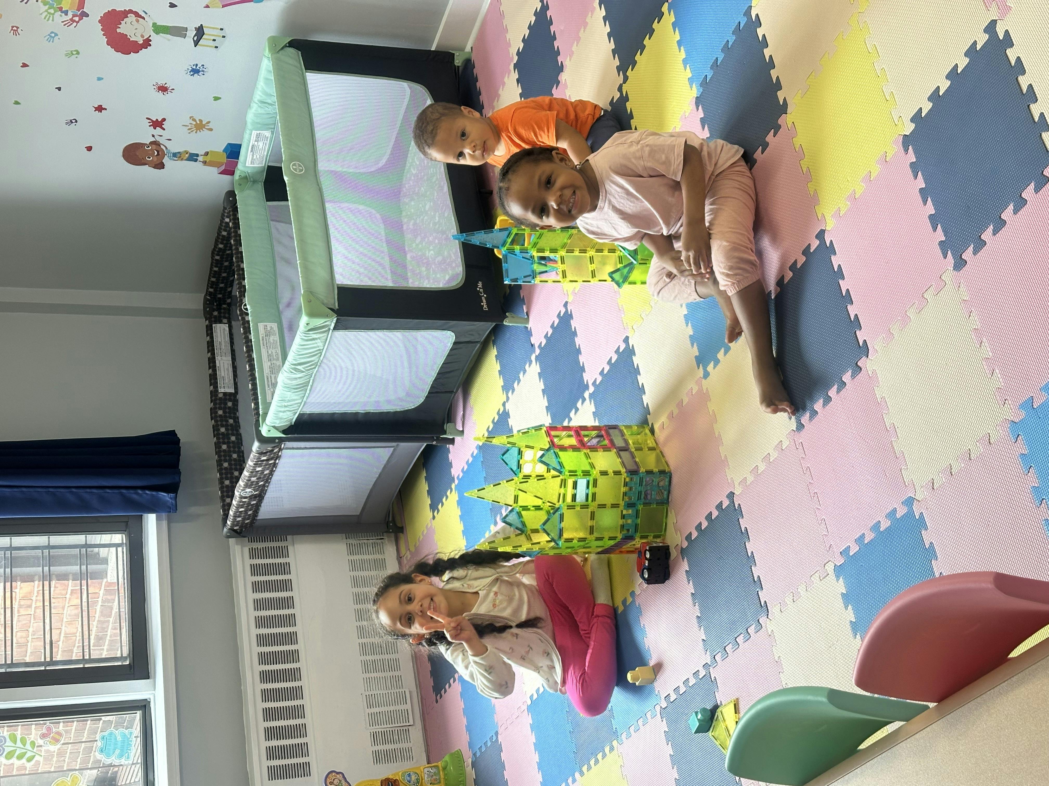 Three children on a colorful puzzle mat in a room with two playpens