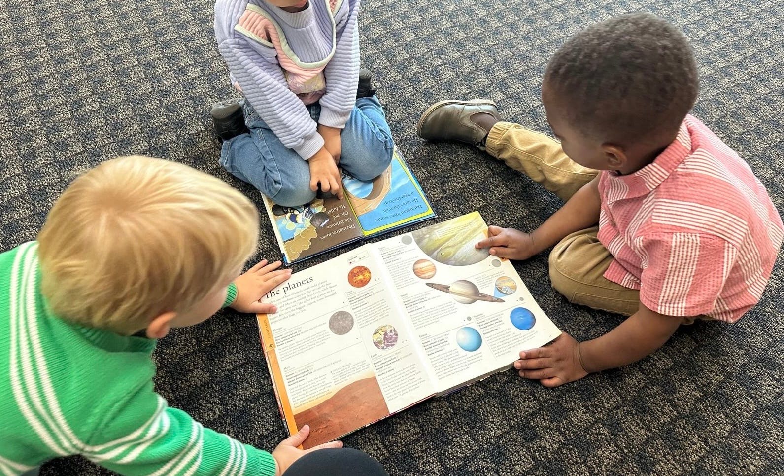 Three children sitting on the floor reading books about planets