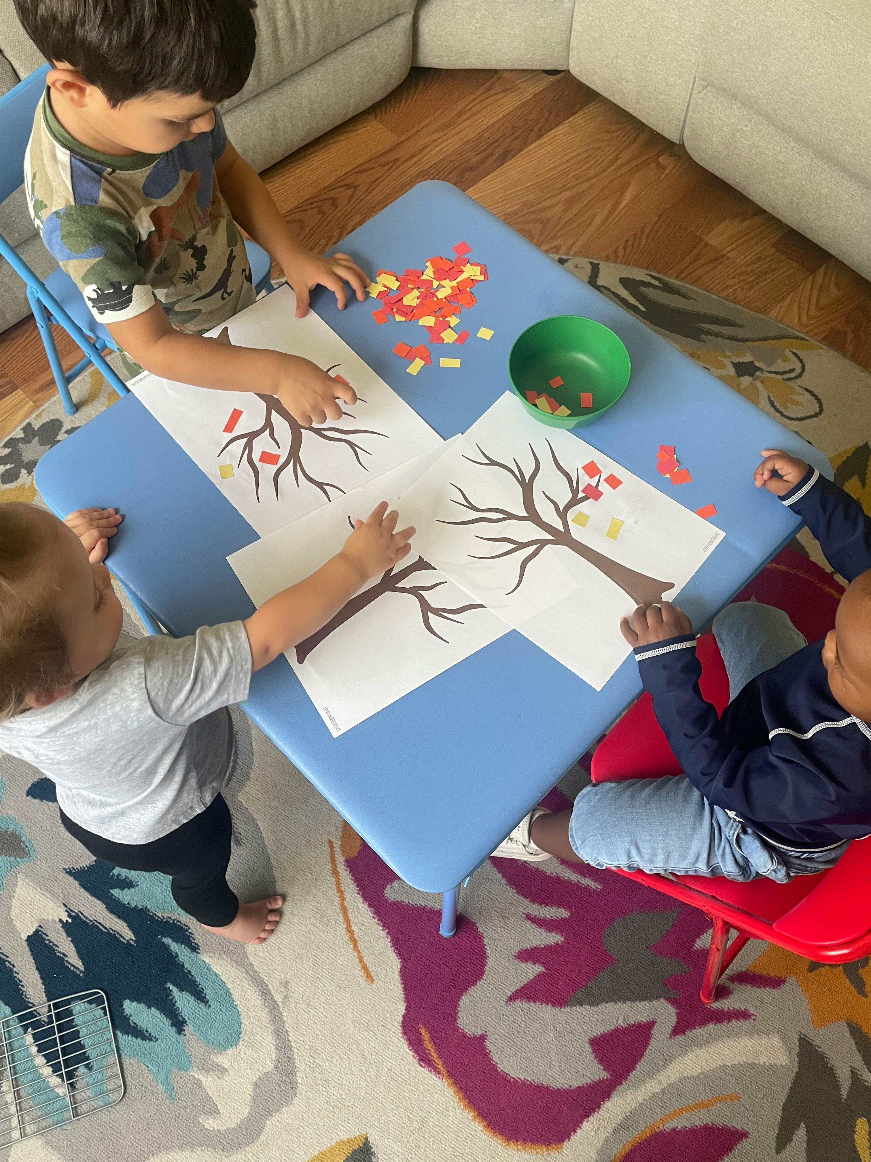 Three children doing a craft activity with paper trees and small paper pieces