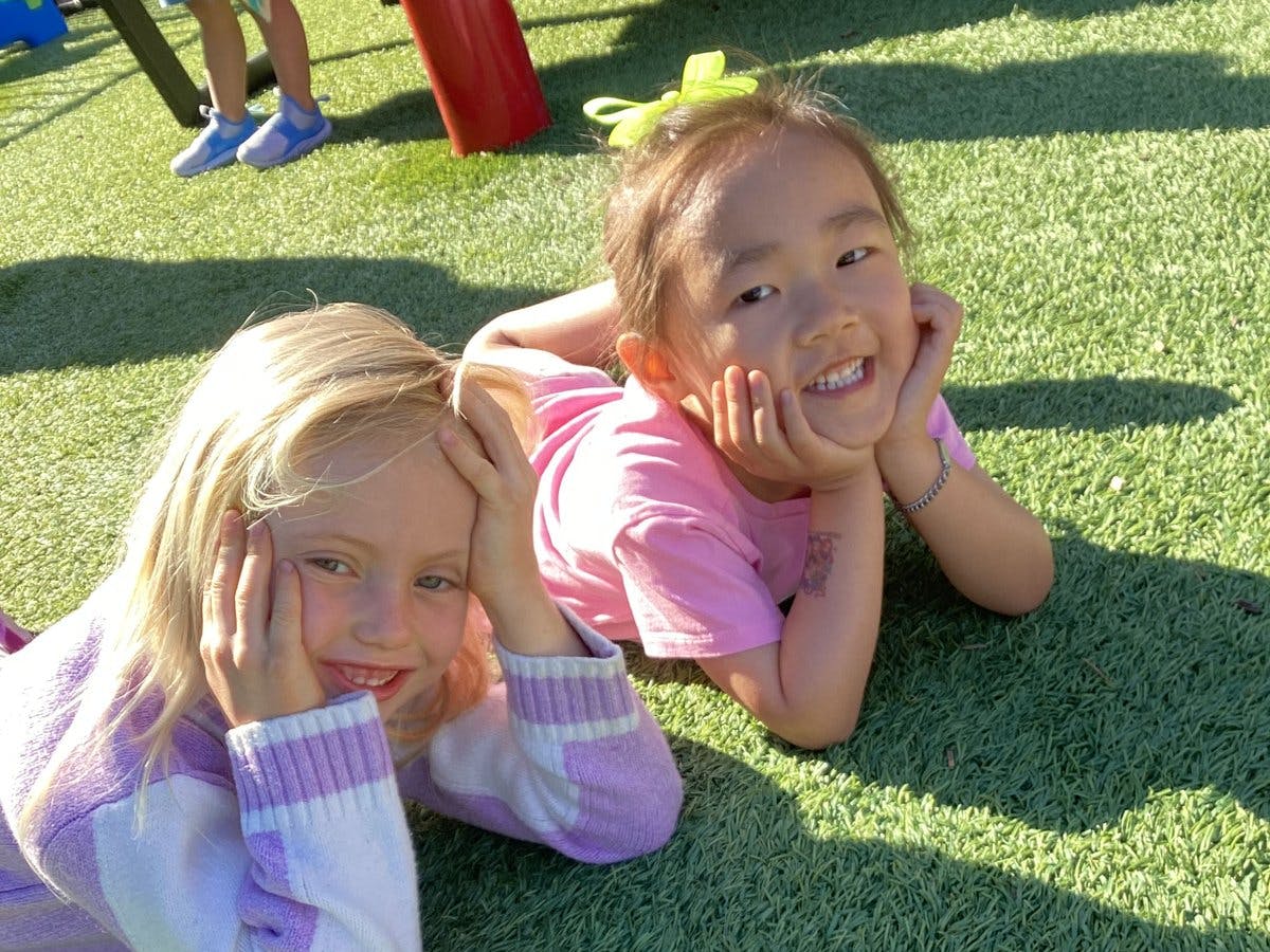 Two children lying on green turf, smiling with hands framing their faces