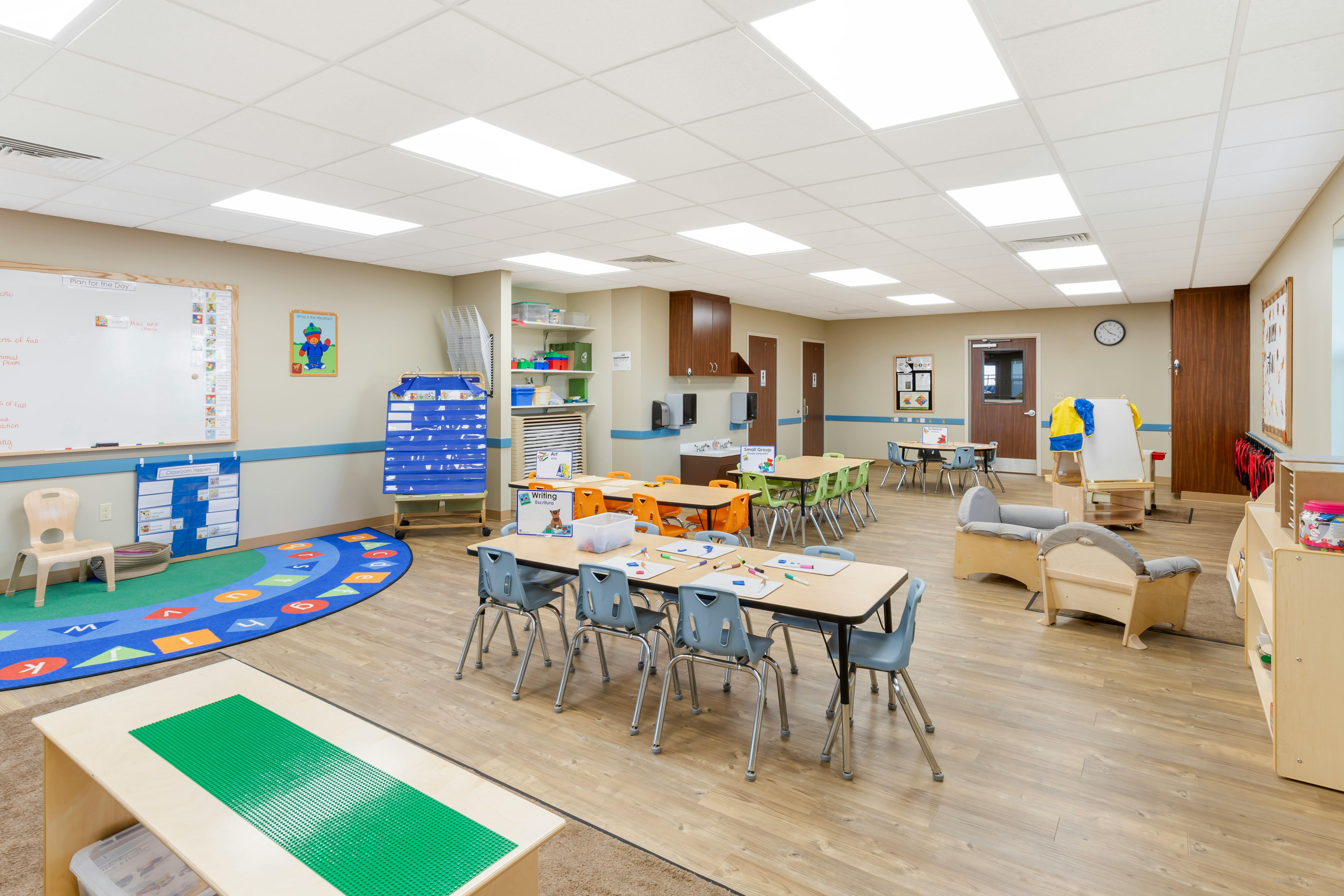 Empty classroom with small tables and chairs, whiteboard, and learning rug