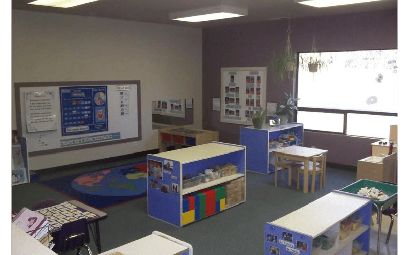 Classroom with whiteboard, colorful rug, shelves with toys, and small tables and chairs