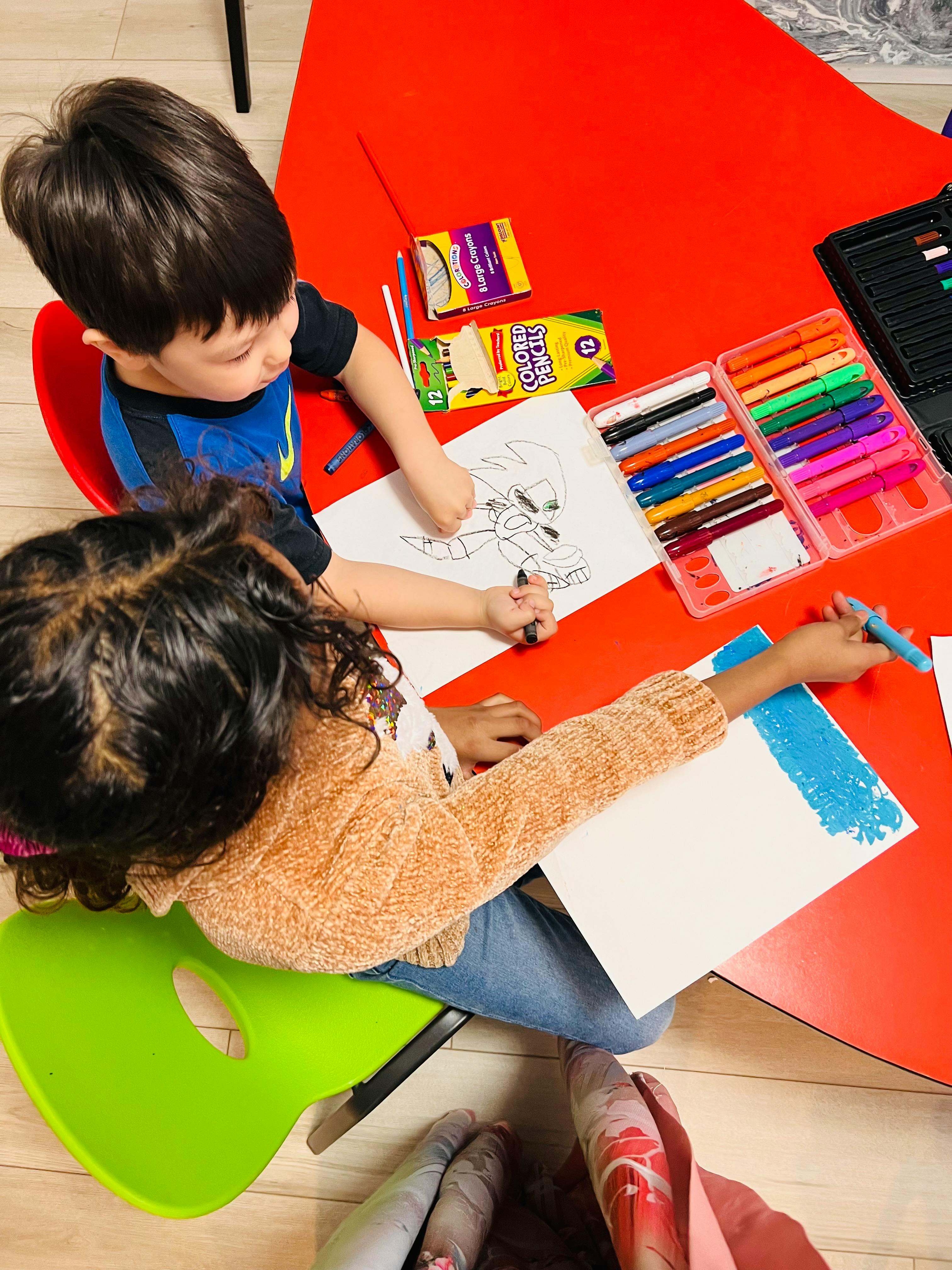 Two children drawing at a red table with markers