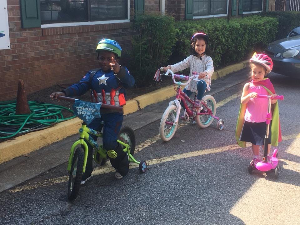 Three children with helmets on a bike, a bike with training wheels, and a scooter