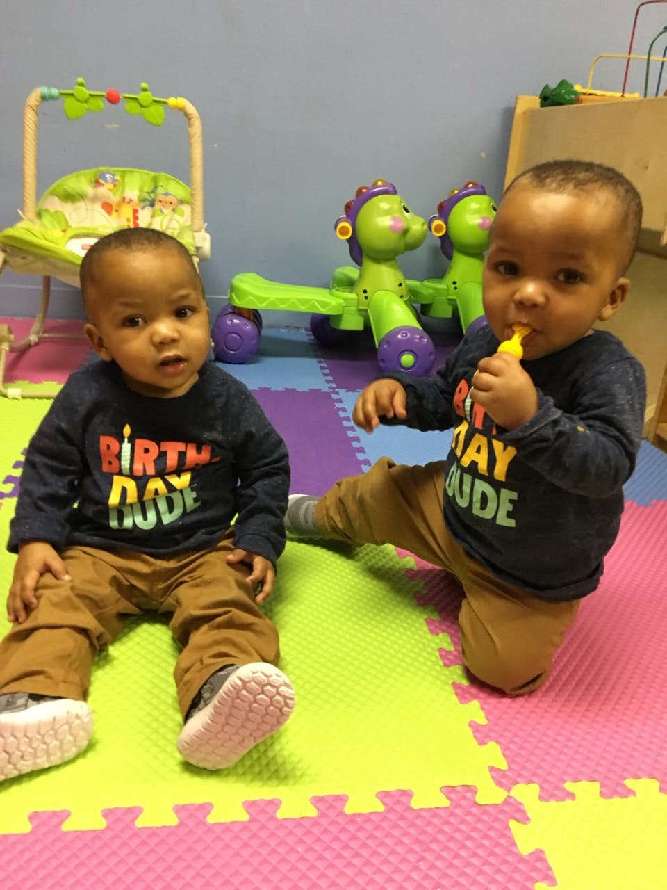 Two children on a colorful mat, one sitting, one kneeling and holding a toy
