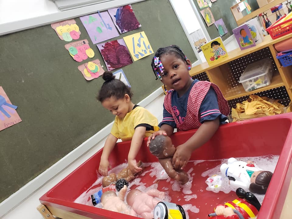 Two children playing with dolls in a tub of soapy water in a classroom