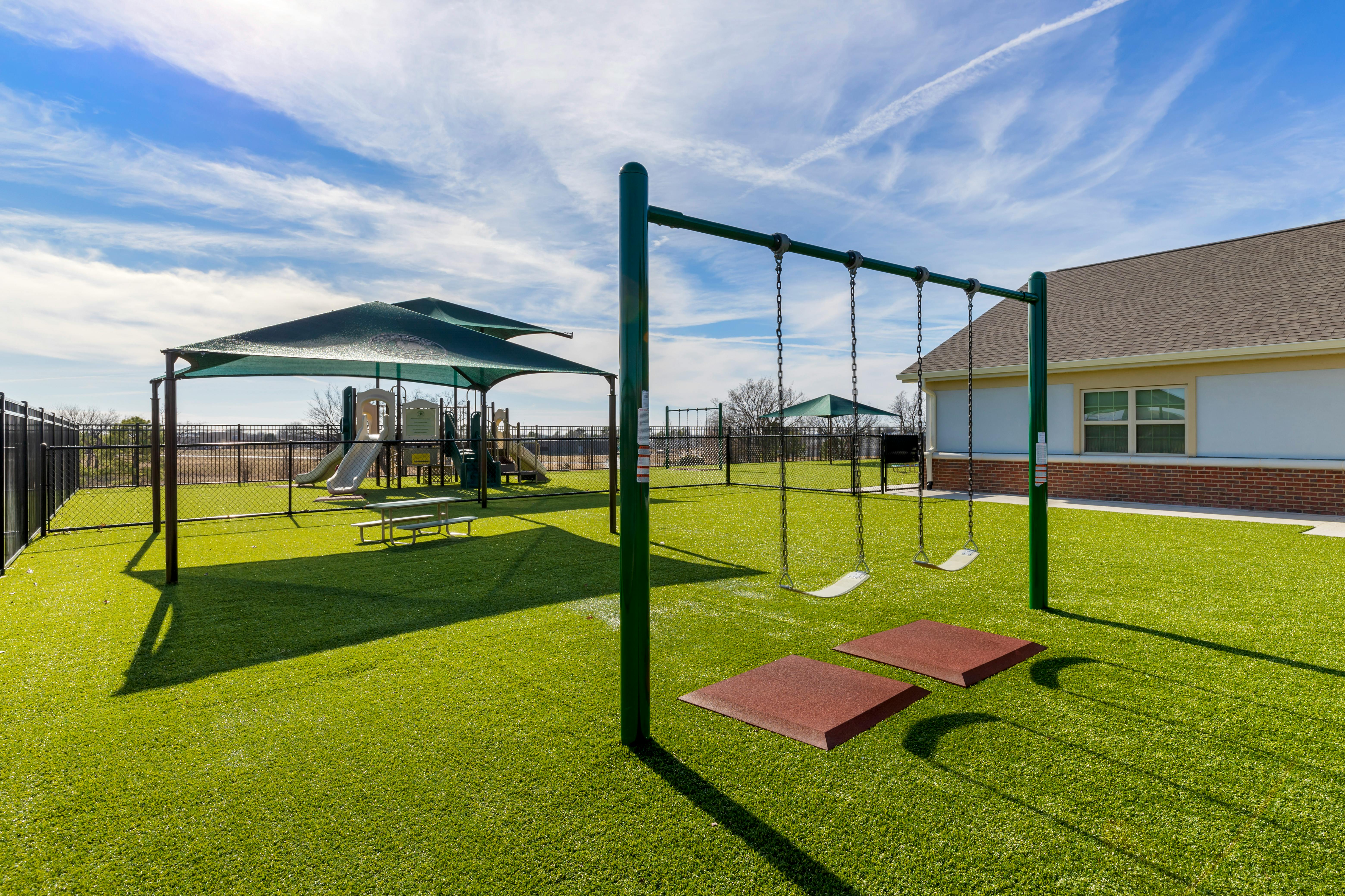 Swingset on artificial grass with a playground and building in the background