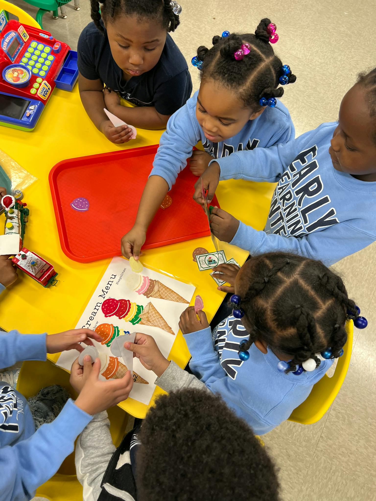 Children playing with an ice cream menu and toy money at a yellow table