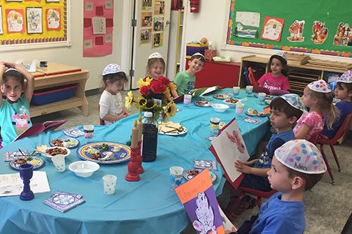Children wearing yarmulkes sitting around a table with food and books