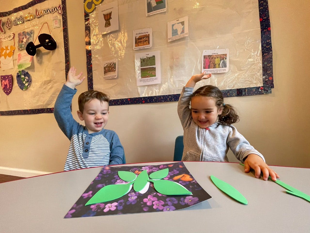 Two children raising their hands at a table with craft supplies