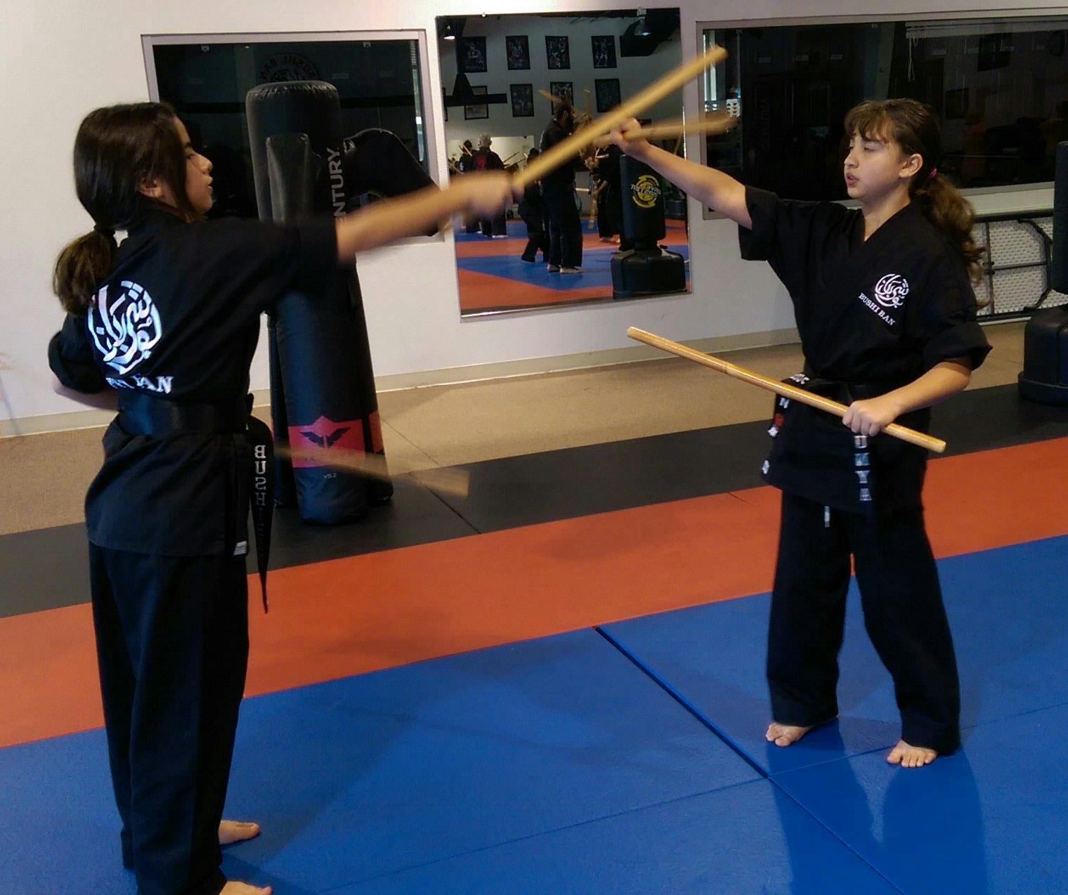 Two children in martial arts uniforms practicing with wooden sticks