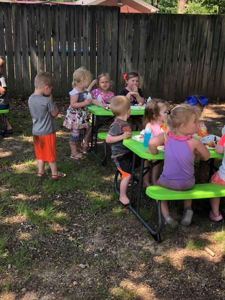 Children sitting and standing around green picnic tables outdoors