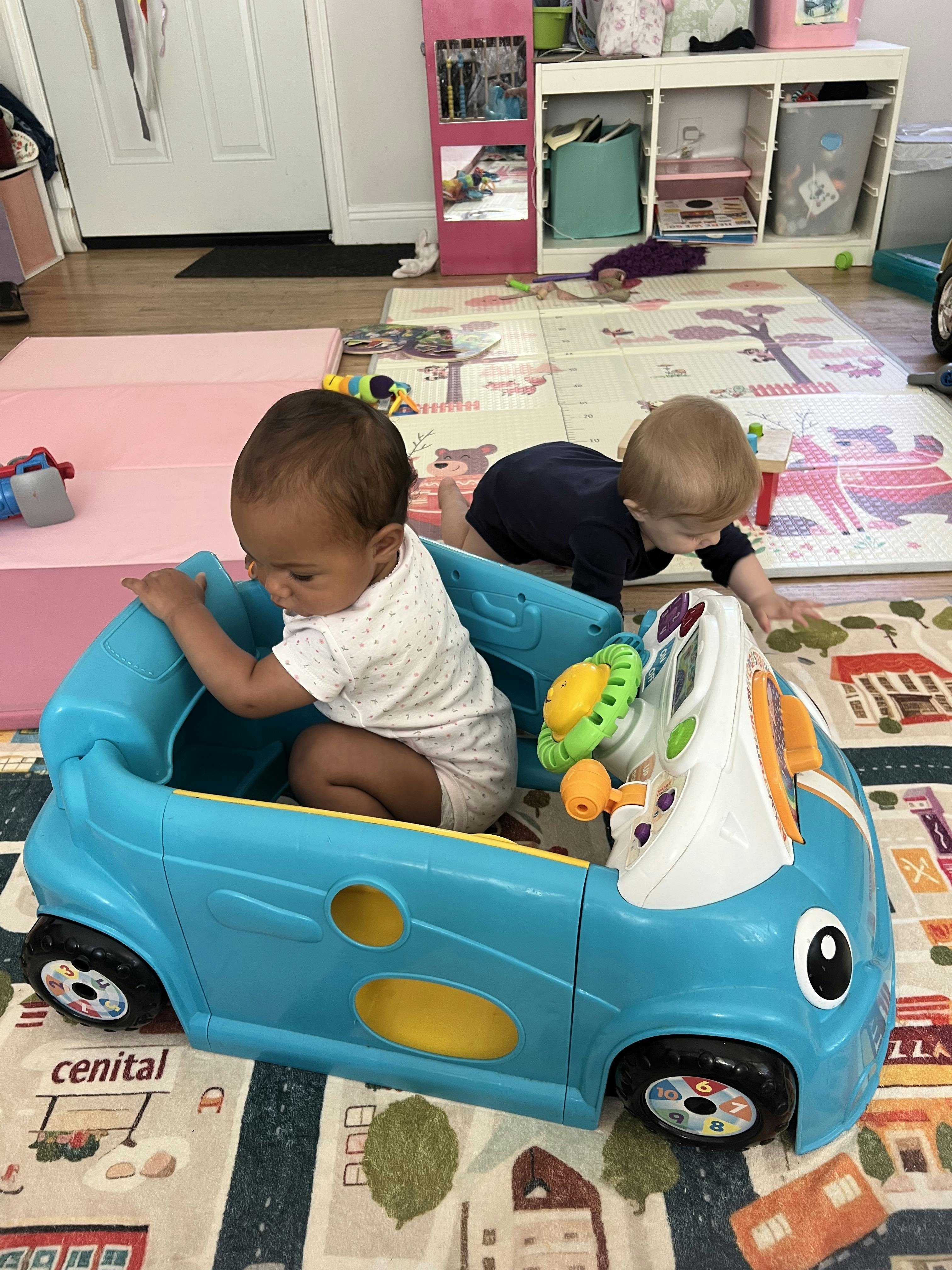 Child sitting in a toy car with another child crawling nearby