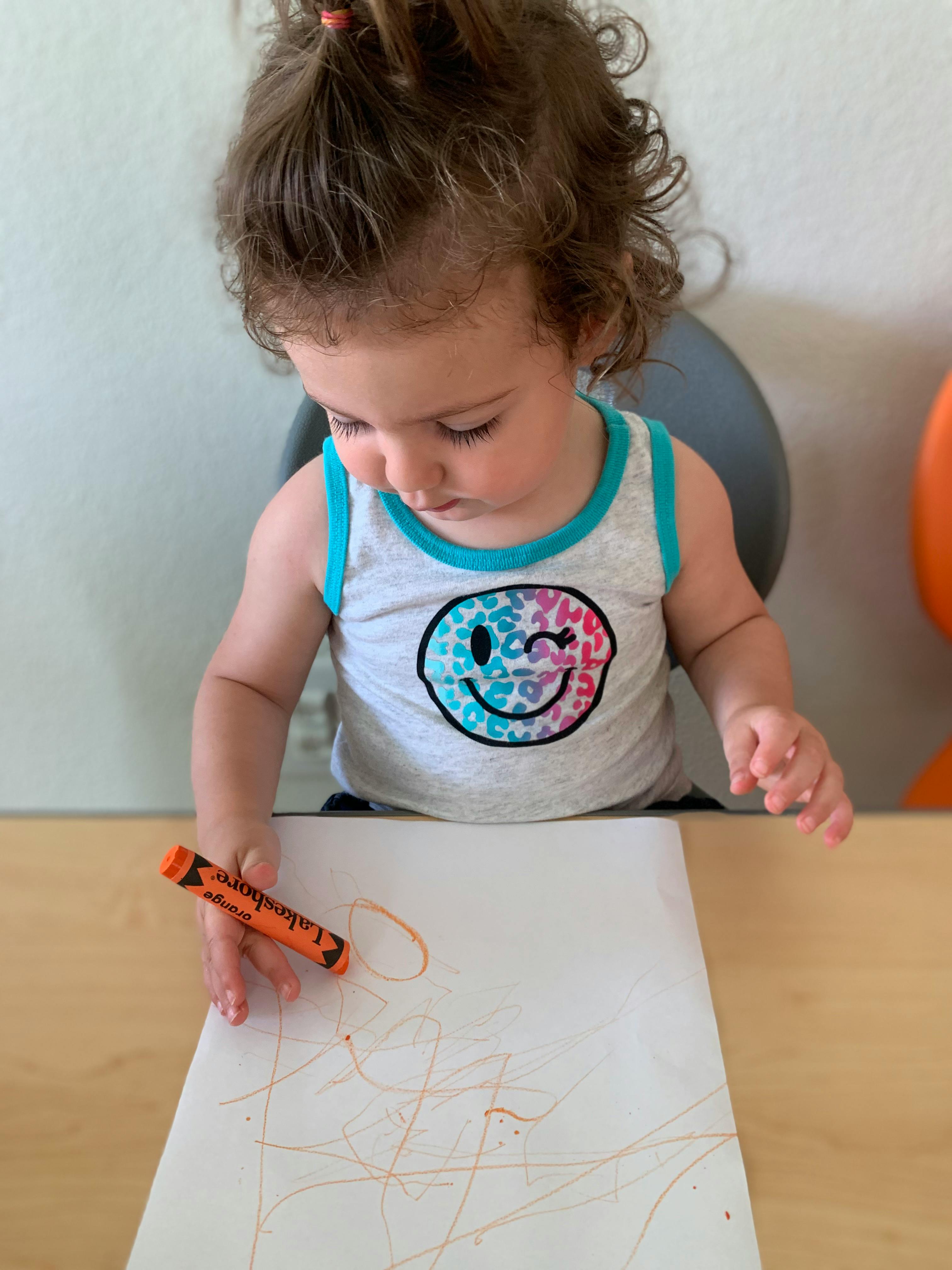 Child drawing on paper with an orange crayon