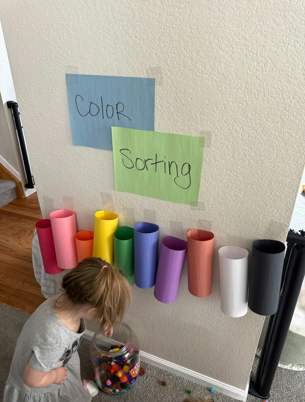 Child sorting colorful pom-poms into tubes on a wall labeled "Color Sorting"