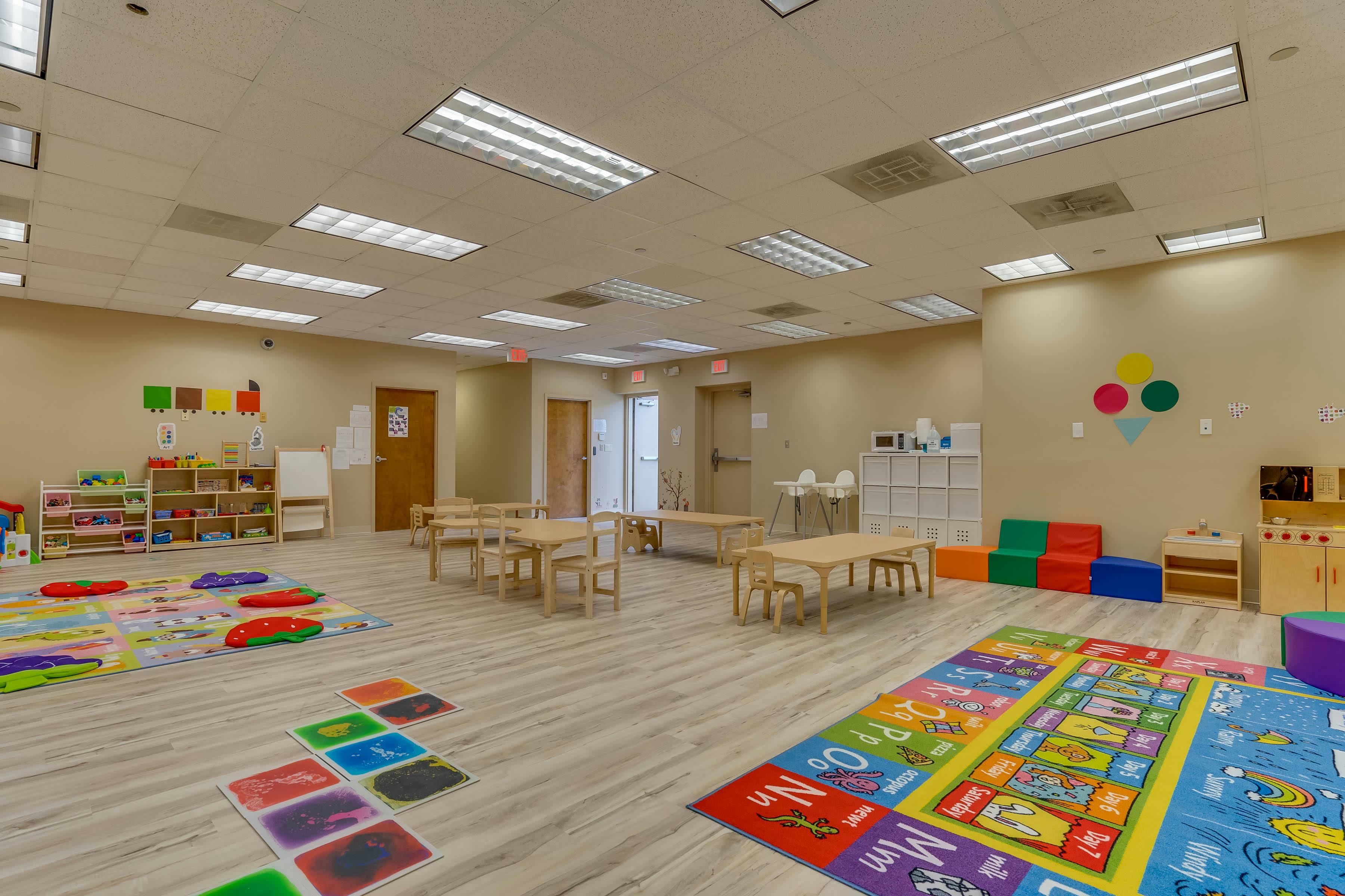 Empty classroom with small tables and chairs, colorful rugs, and shelves