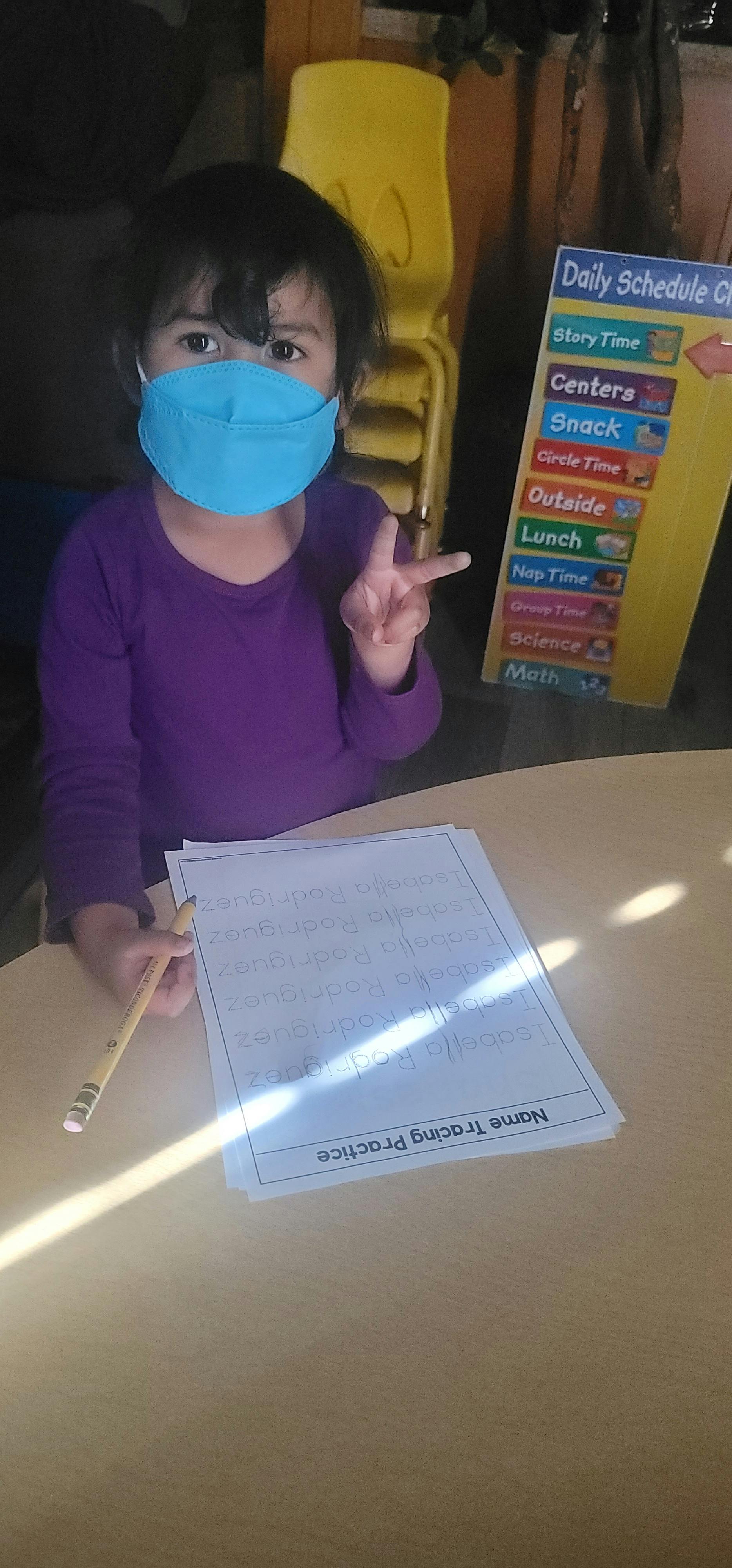 Child in a mask holding a pencil and showing a peace sign