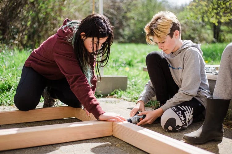 Two teens assembling wooden planks outdoors with a drill