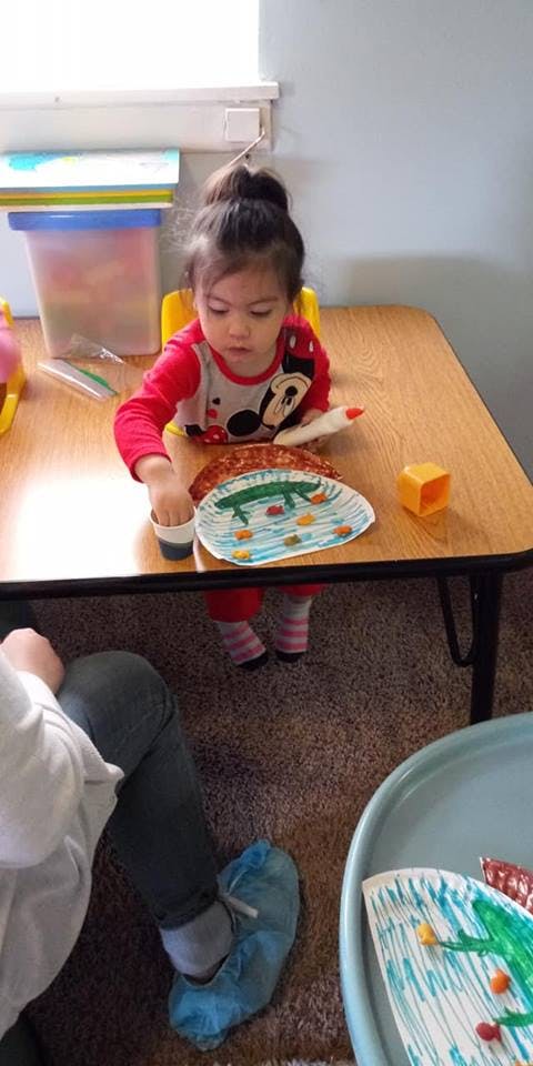 Child sitting at a table, reaching for a cup next to a decorated paper plate