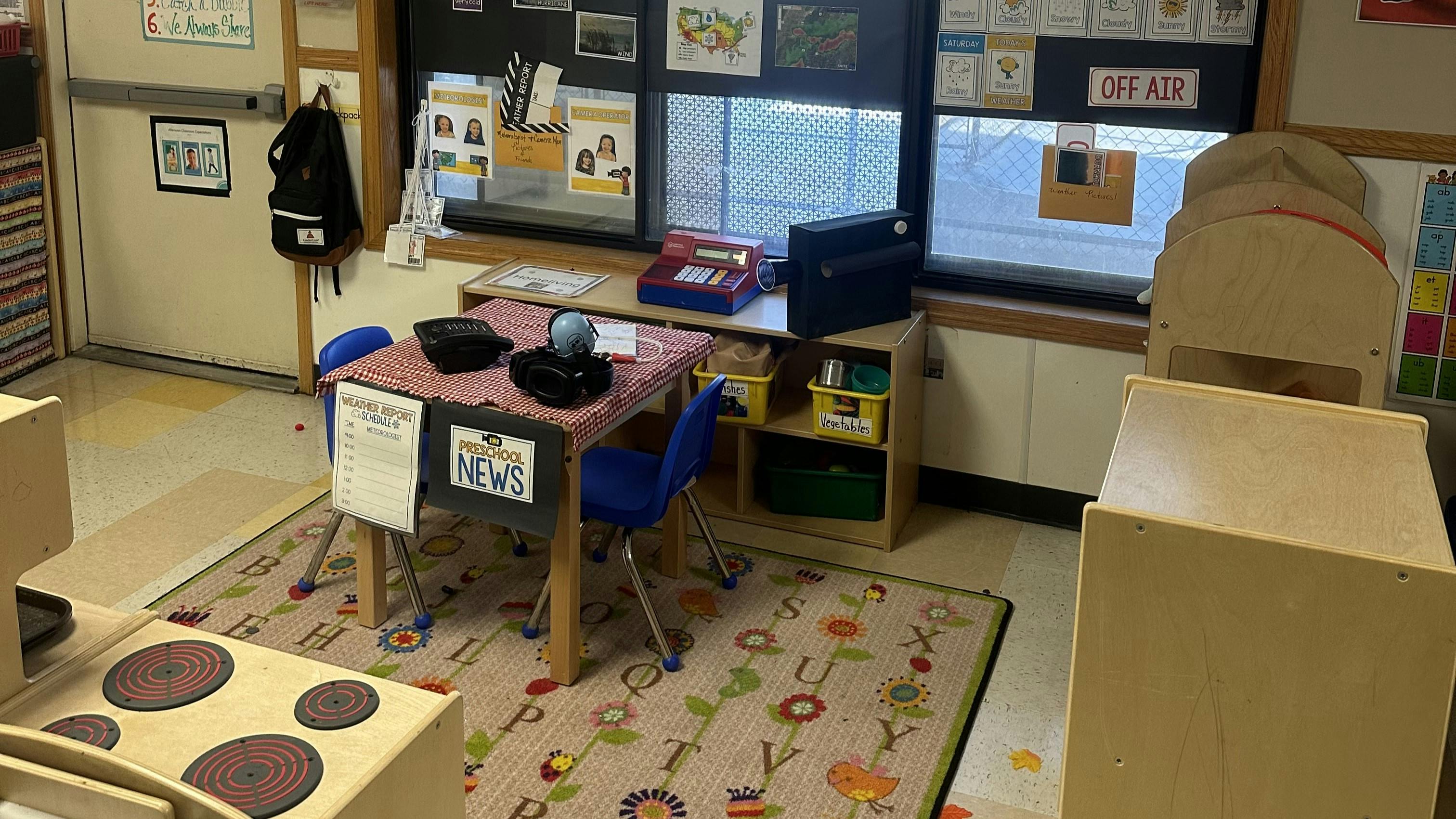 Classroom corner with a small table, chairs, play kitchen, and a window display for pretend news reports