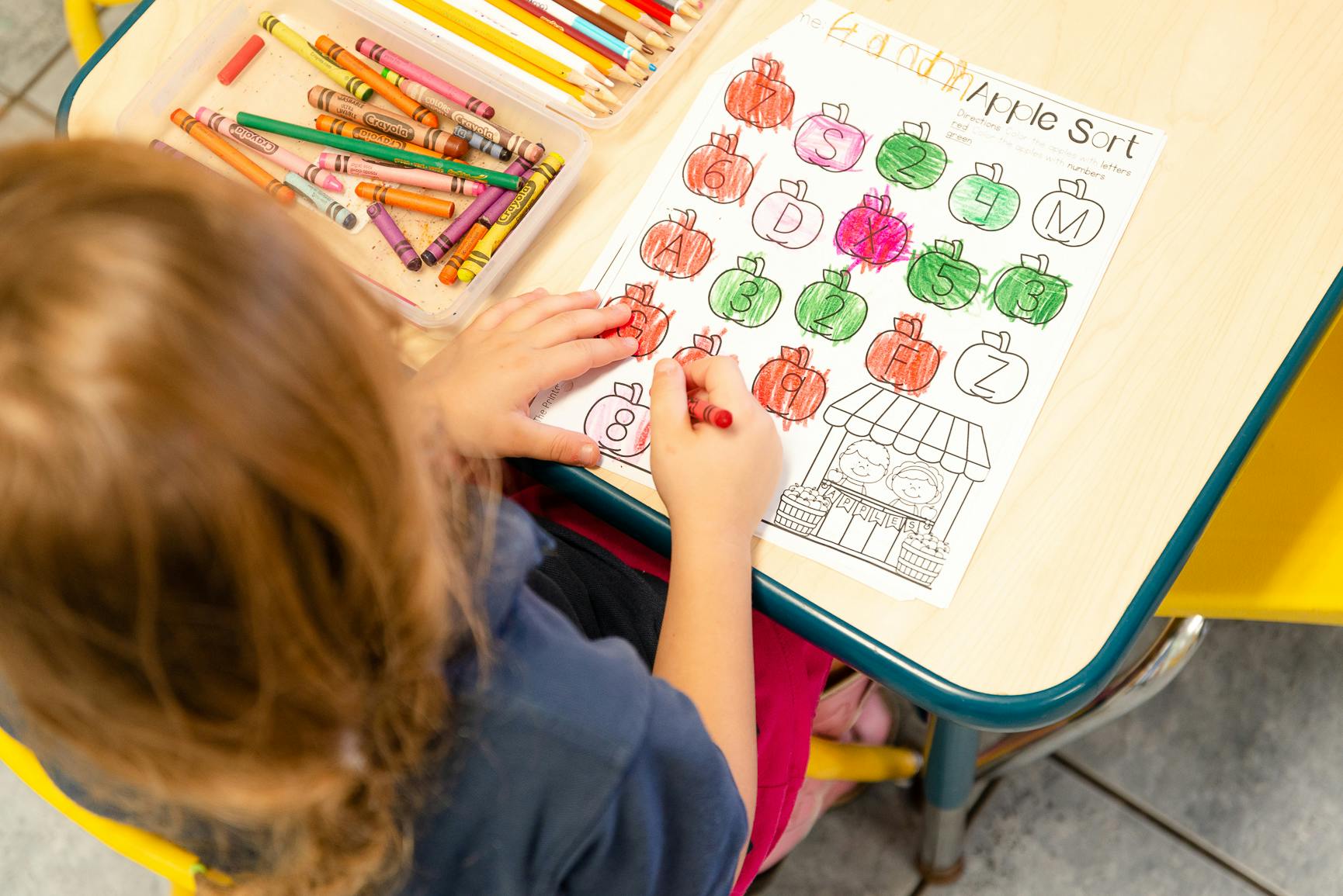 Child coloring a worksheet with apples and letters at a desk with crayons