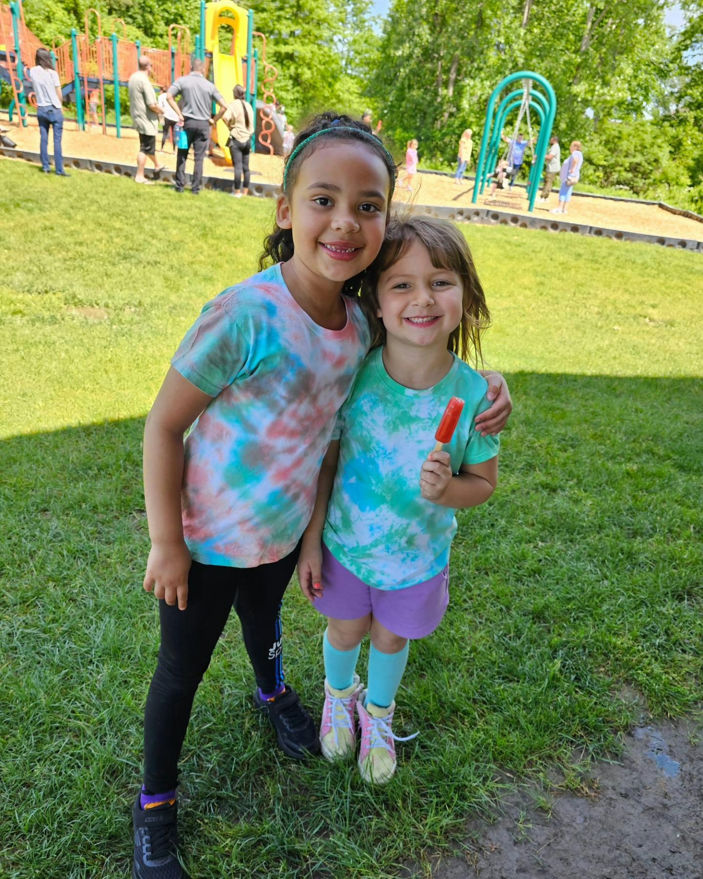 Two children in tie-dye shirts standing together, one holding a popsicle