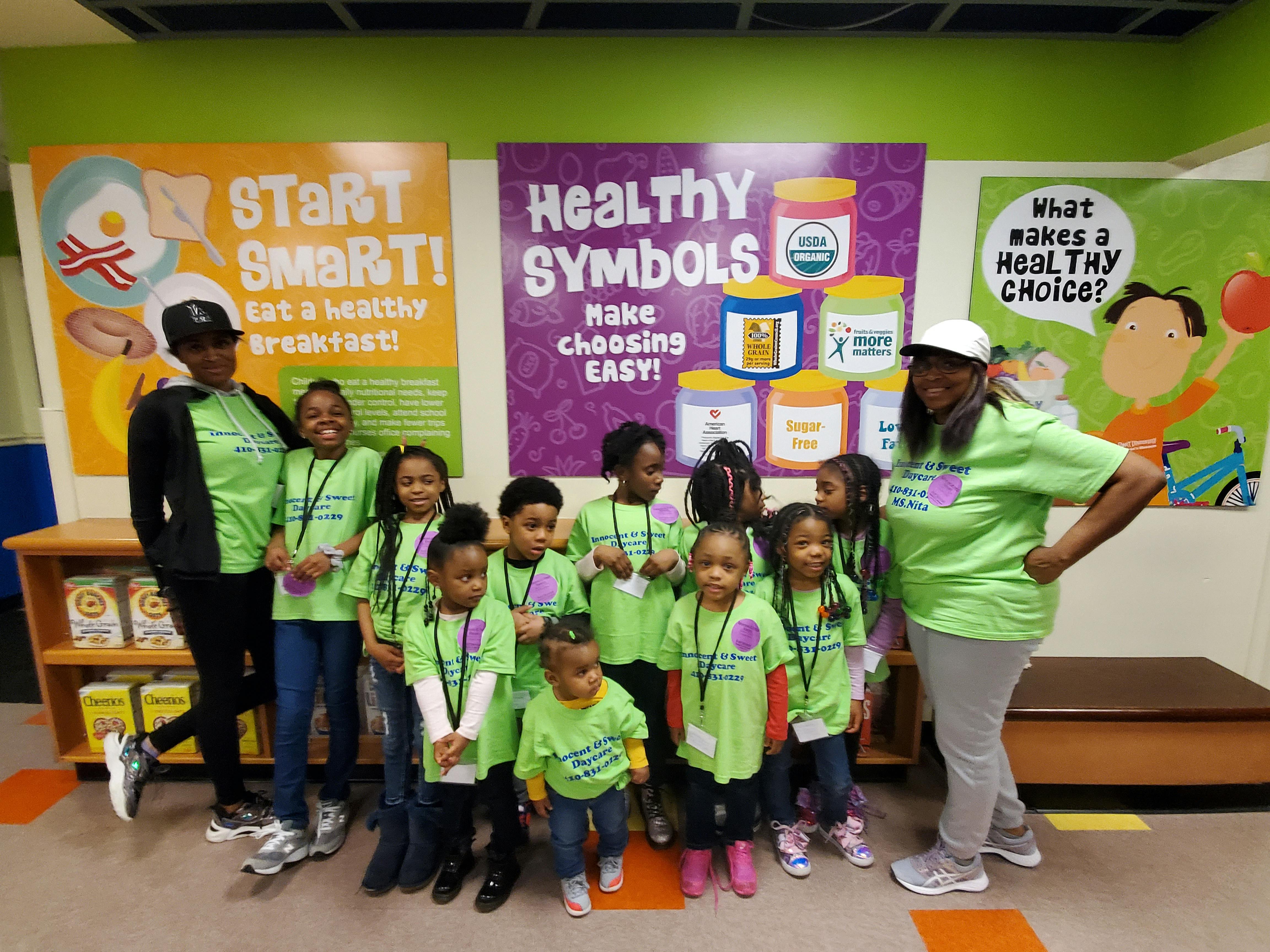 Two adults and multiple children in green shirts standing in front of posters