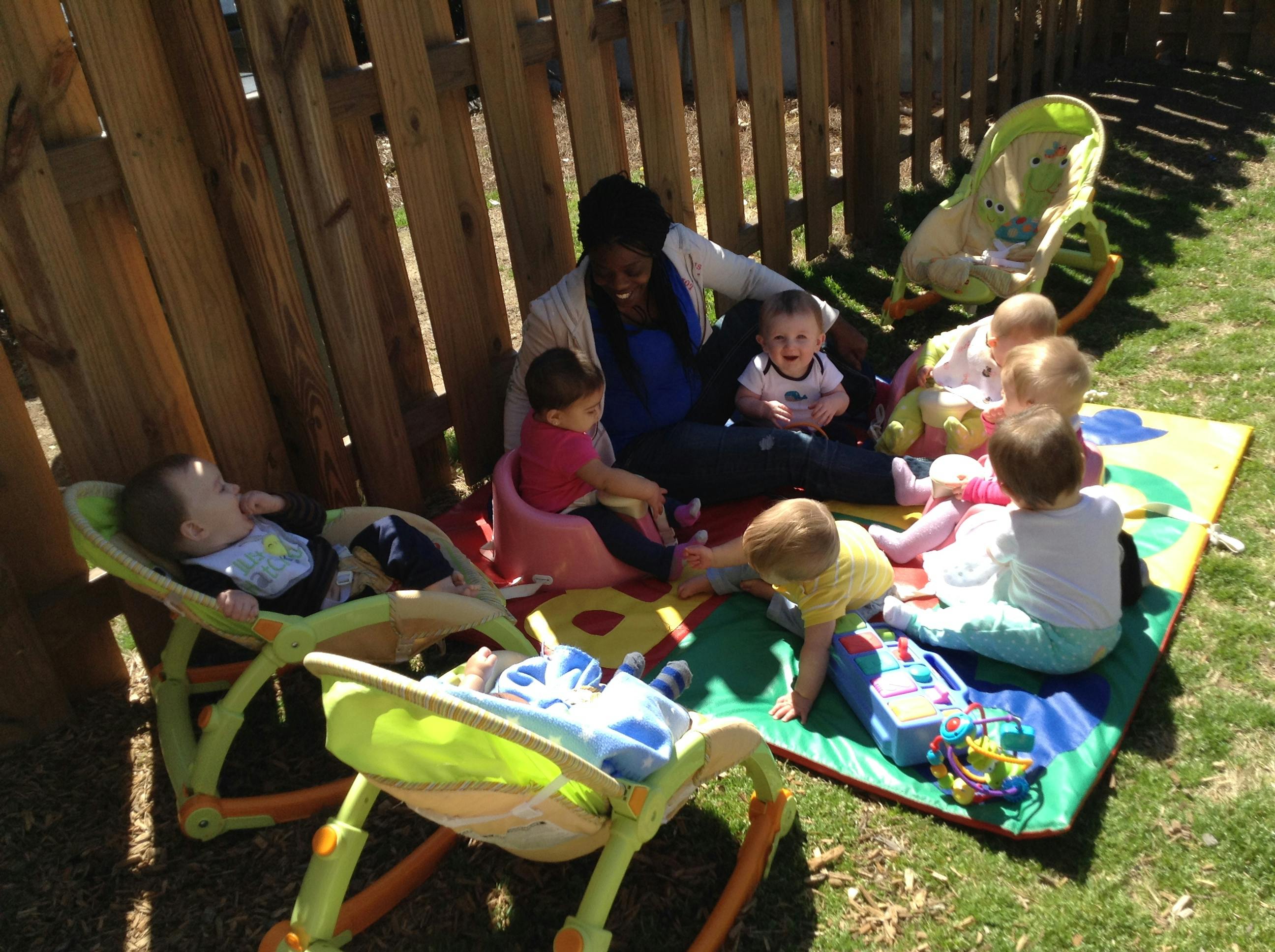 Adult sitting on a mat outdoors surrounded by several children and infant toys