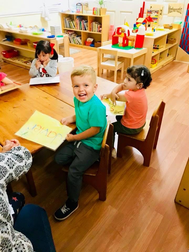Three children at a table in a classroom, one holding up a drawing