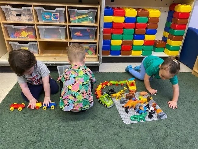 Three children playing with toys on a green carpet in a classroom