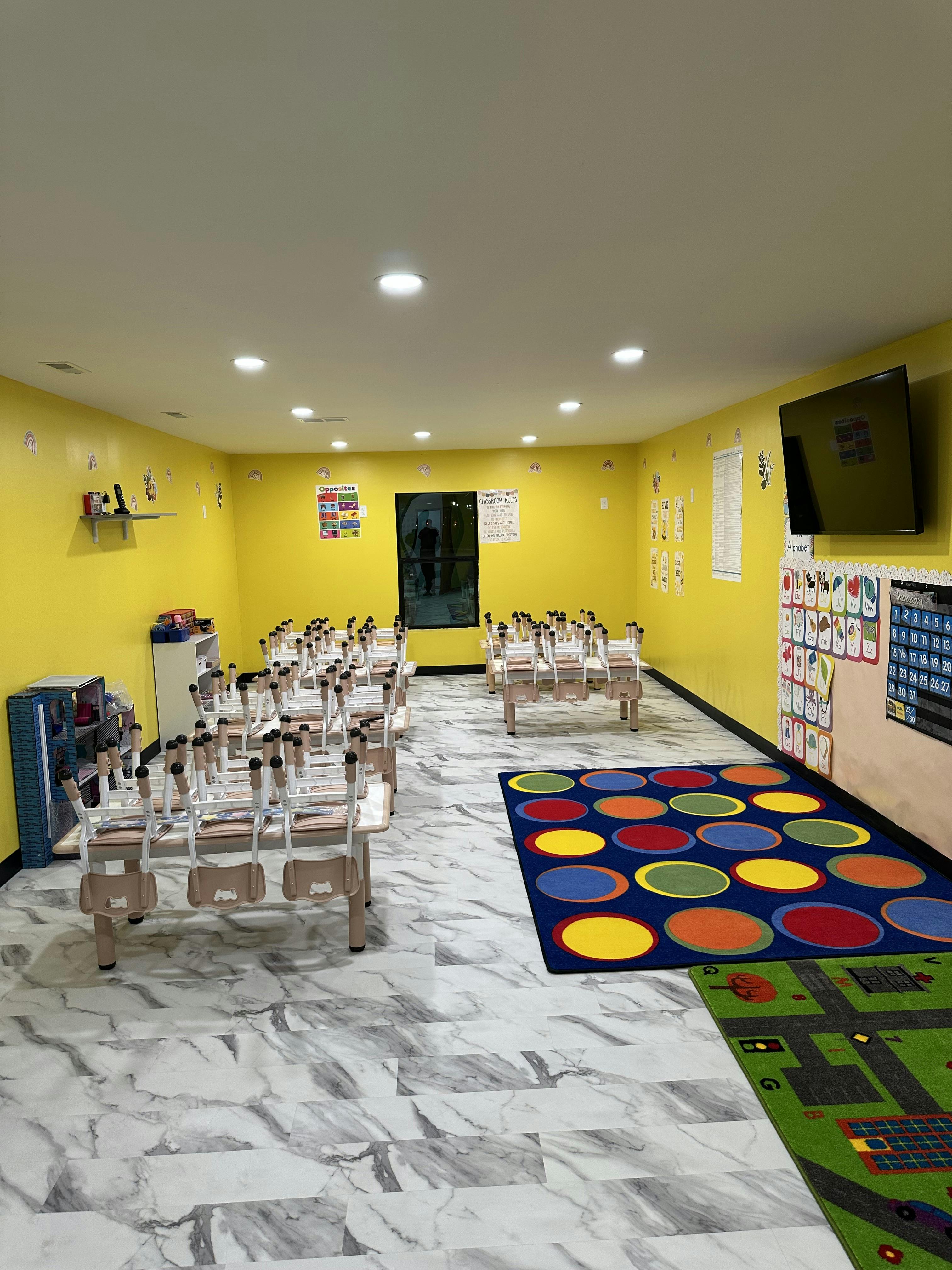 Classroom with small tables and chairs, colorful rugs, and yellow walls