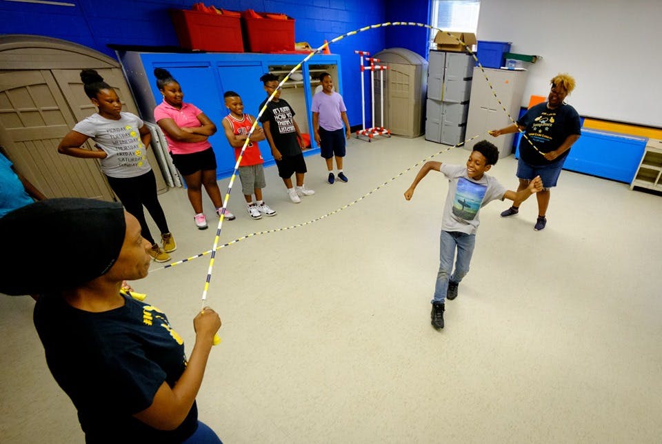 People jump roping with a long rope in a room with other people watching