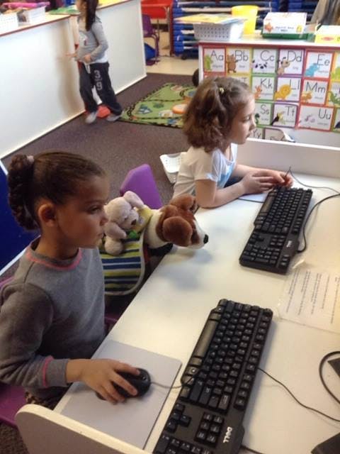 Two children at desks with keyboards and a mouse, one child standing in background