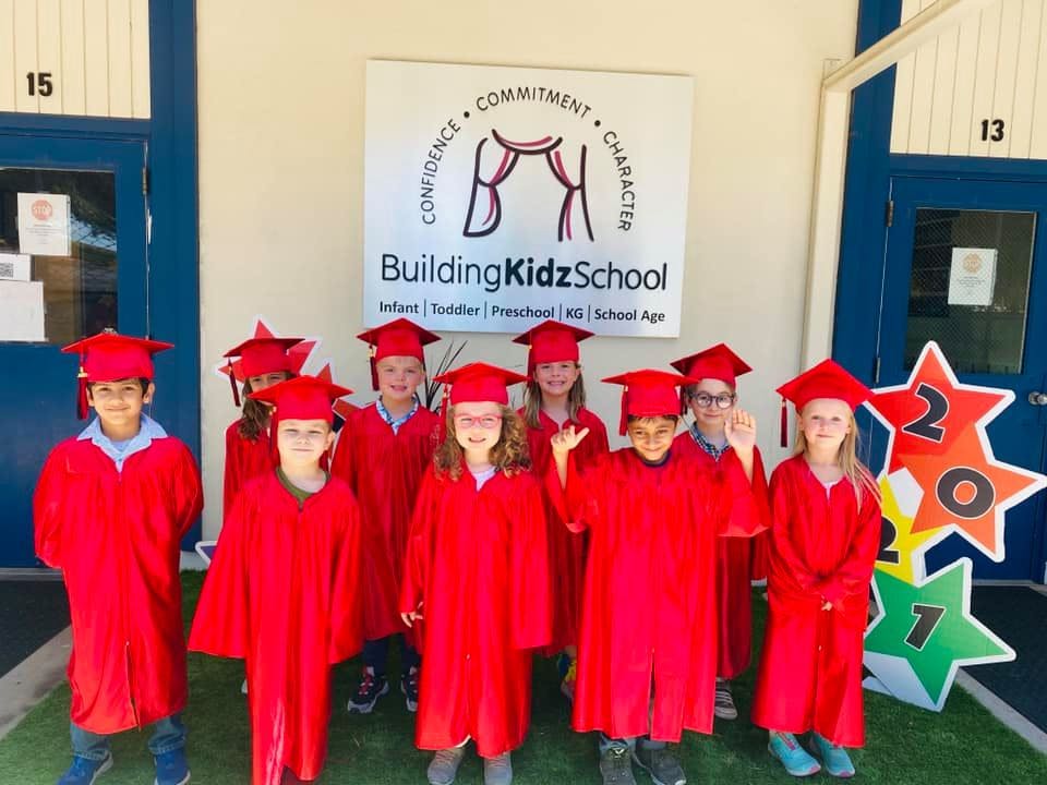 Children in red graduation caps and gowns standing in front of a school sign