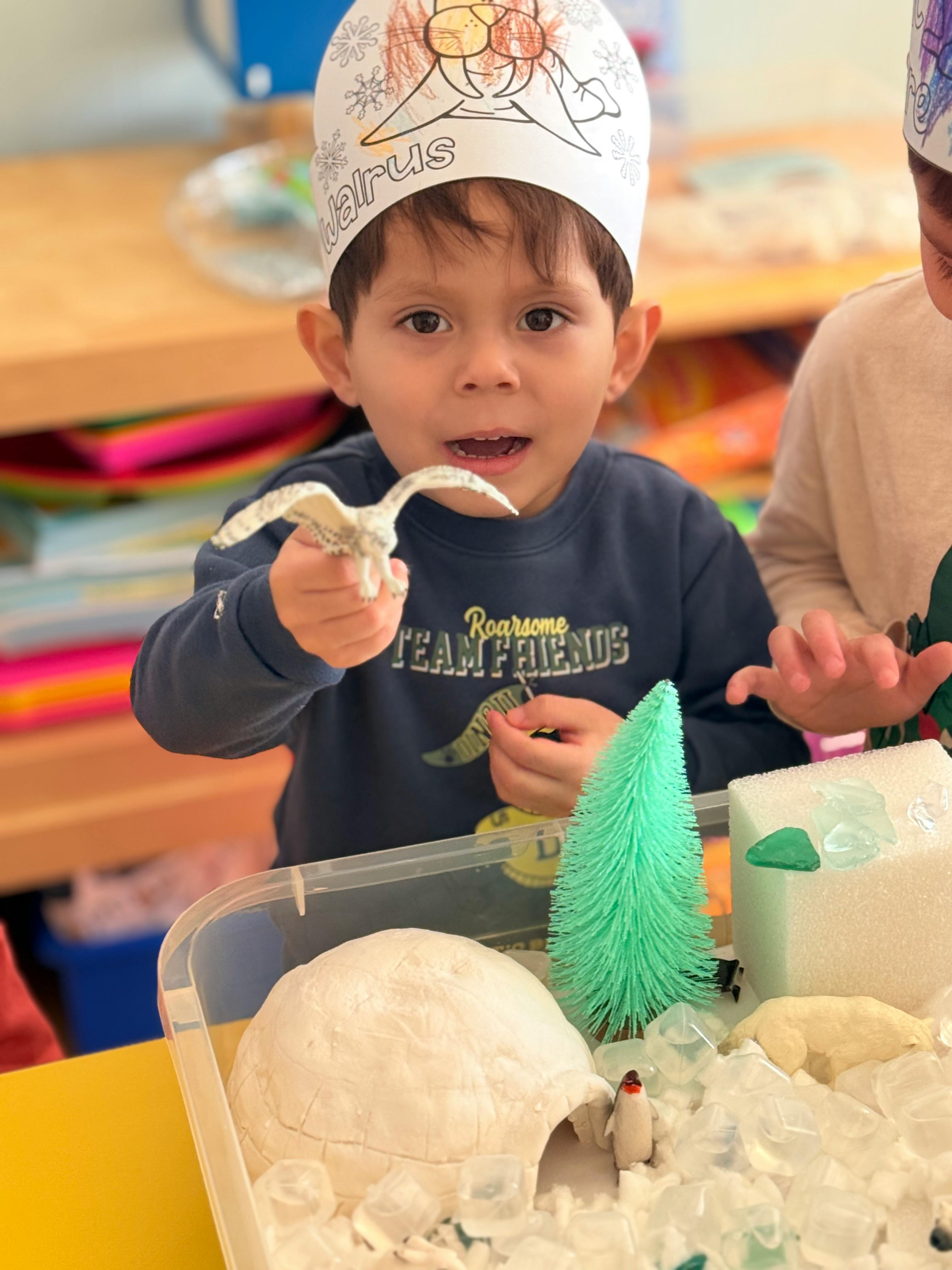 Child in a paper hat holding a toy bird over a sensory bin with an igloo