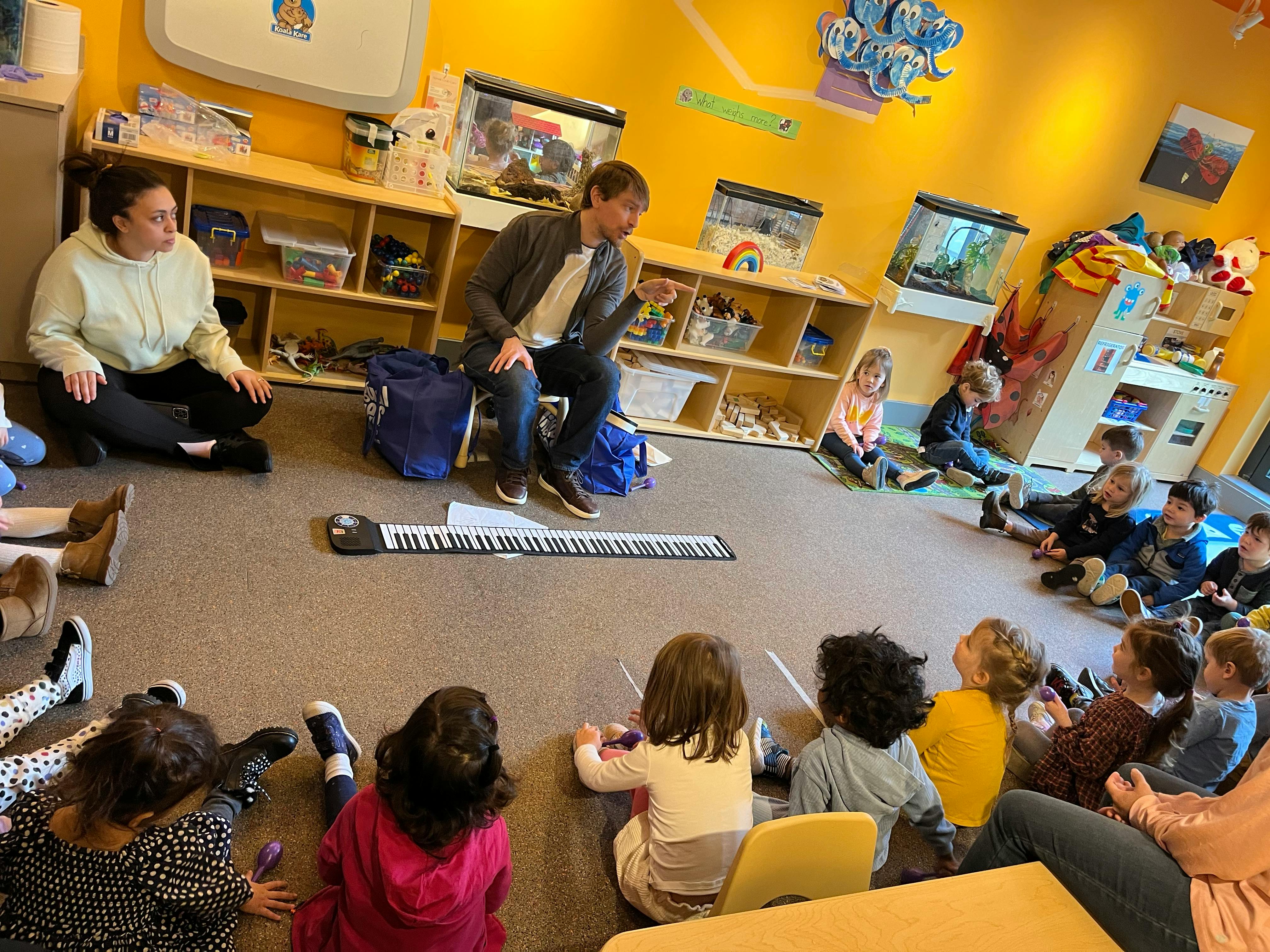 Two adults and multiple children sitting on the floor in a classroom