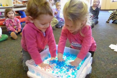 Two children playing with blue and white beads in a clear bin