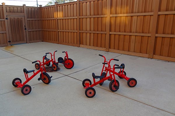 Four red children's tricycles on a concrete surface next to a wooden fence