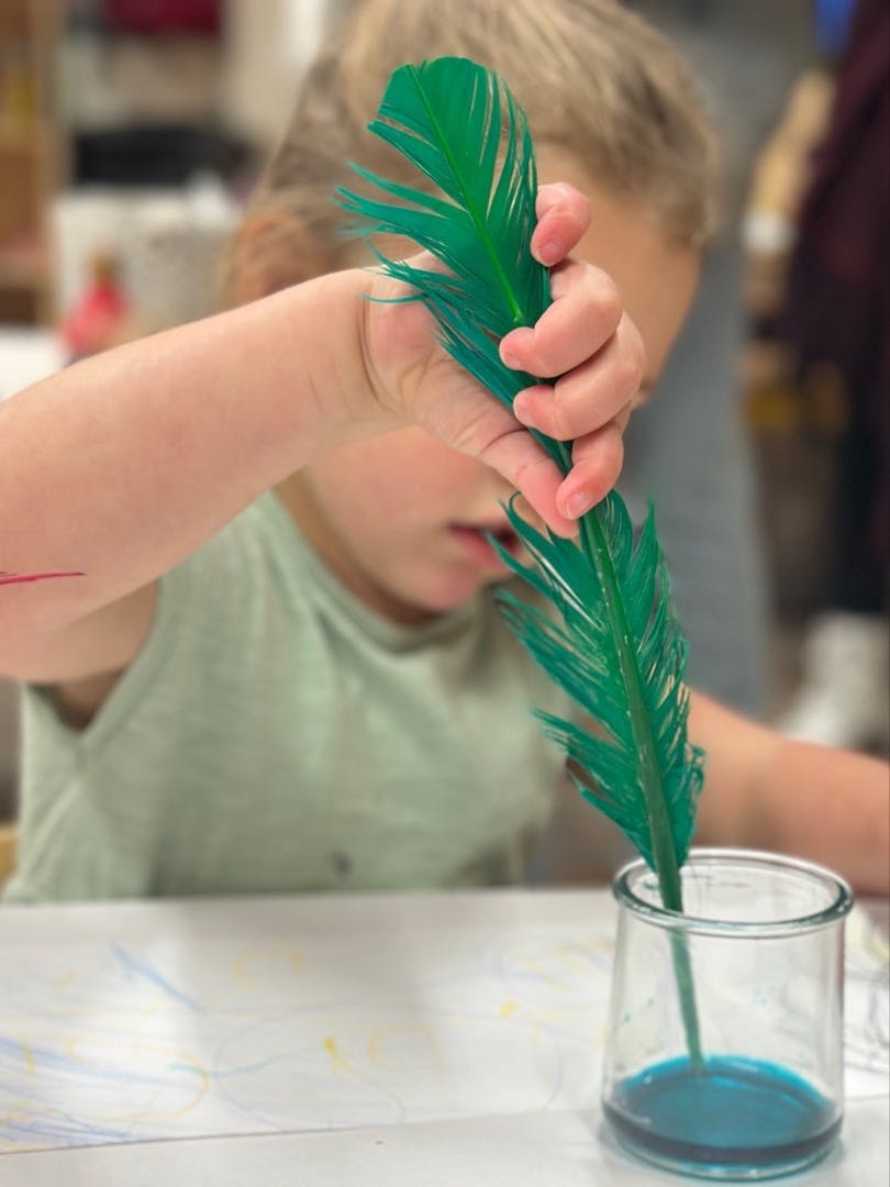 Child holding a green feather over a glass of blue liquid