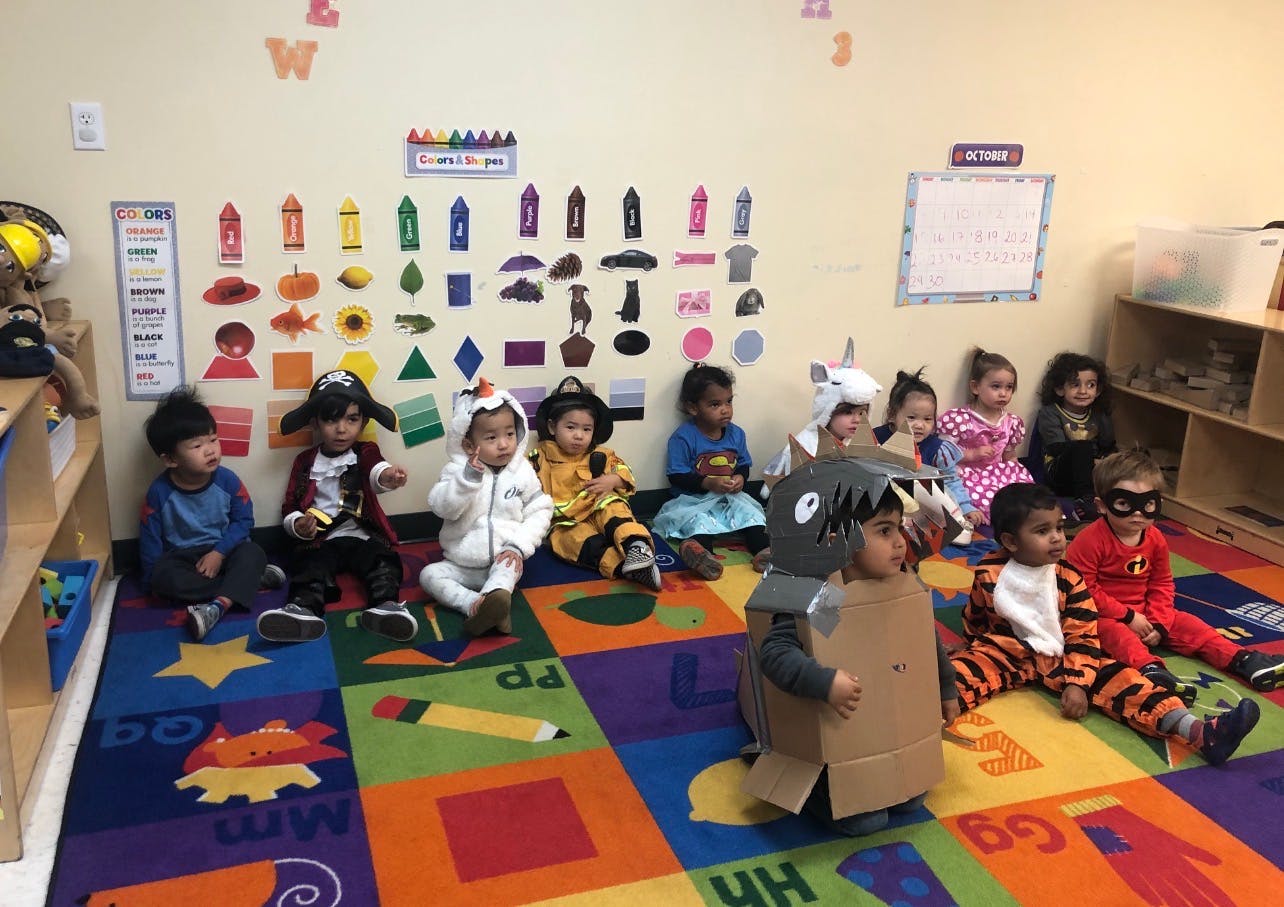 Group of children in various costumes sitting on a colorful rug in a classroom