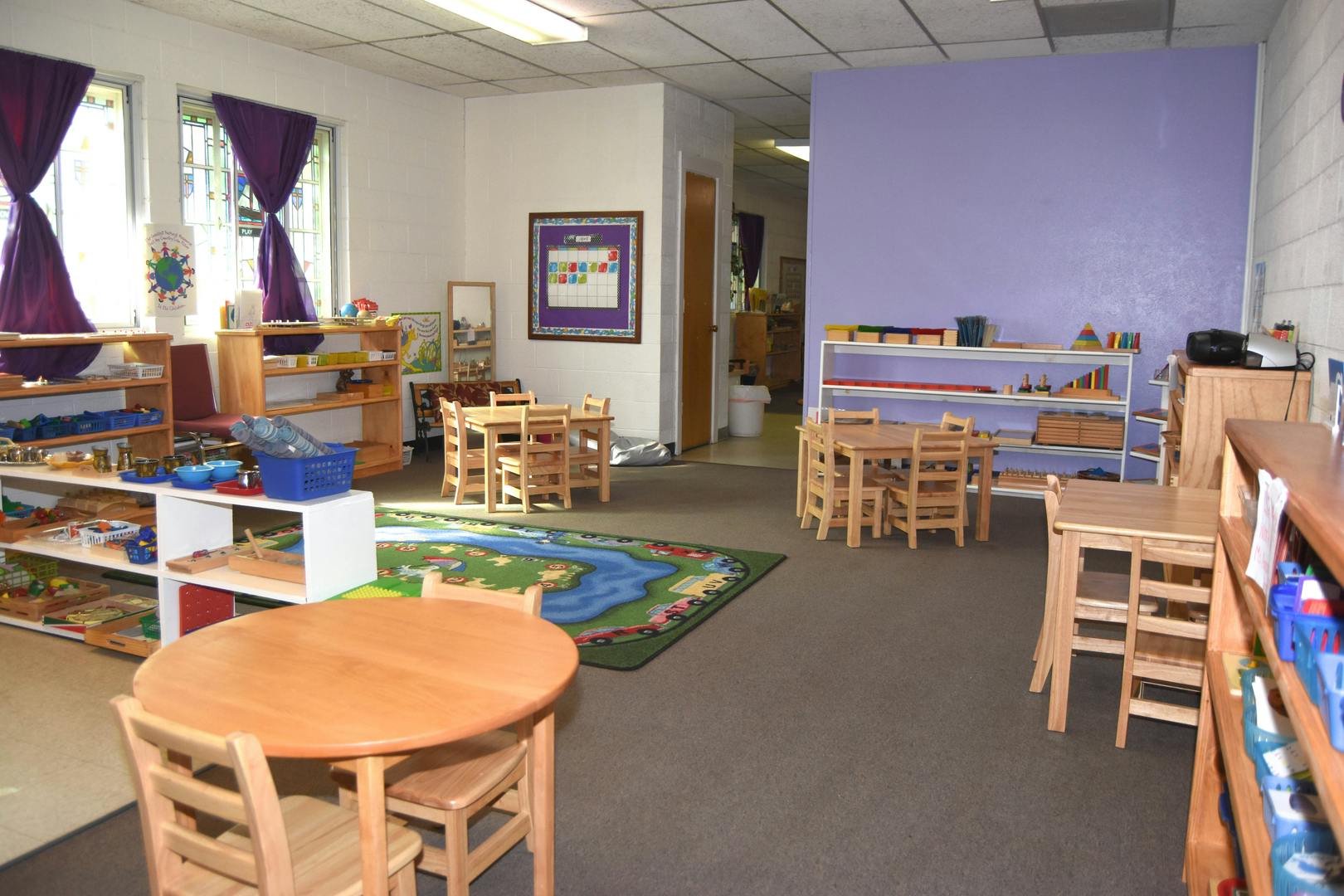 Classroom with small wooden tables and chairs, shelves with learning materials