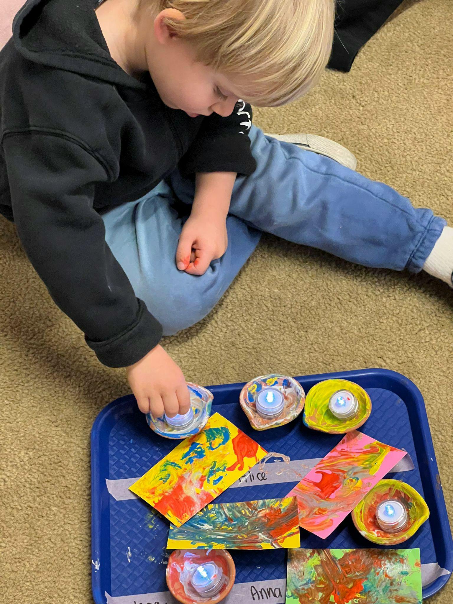 Child reaching for a small painted bowl on a blue tray with other crafts