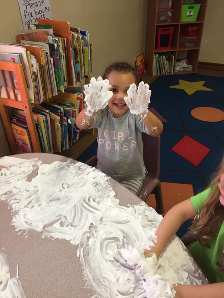 Child with hands covered in white foam, smiling at a table
