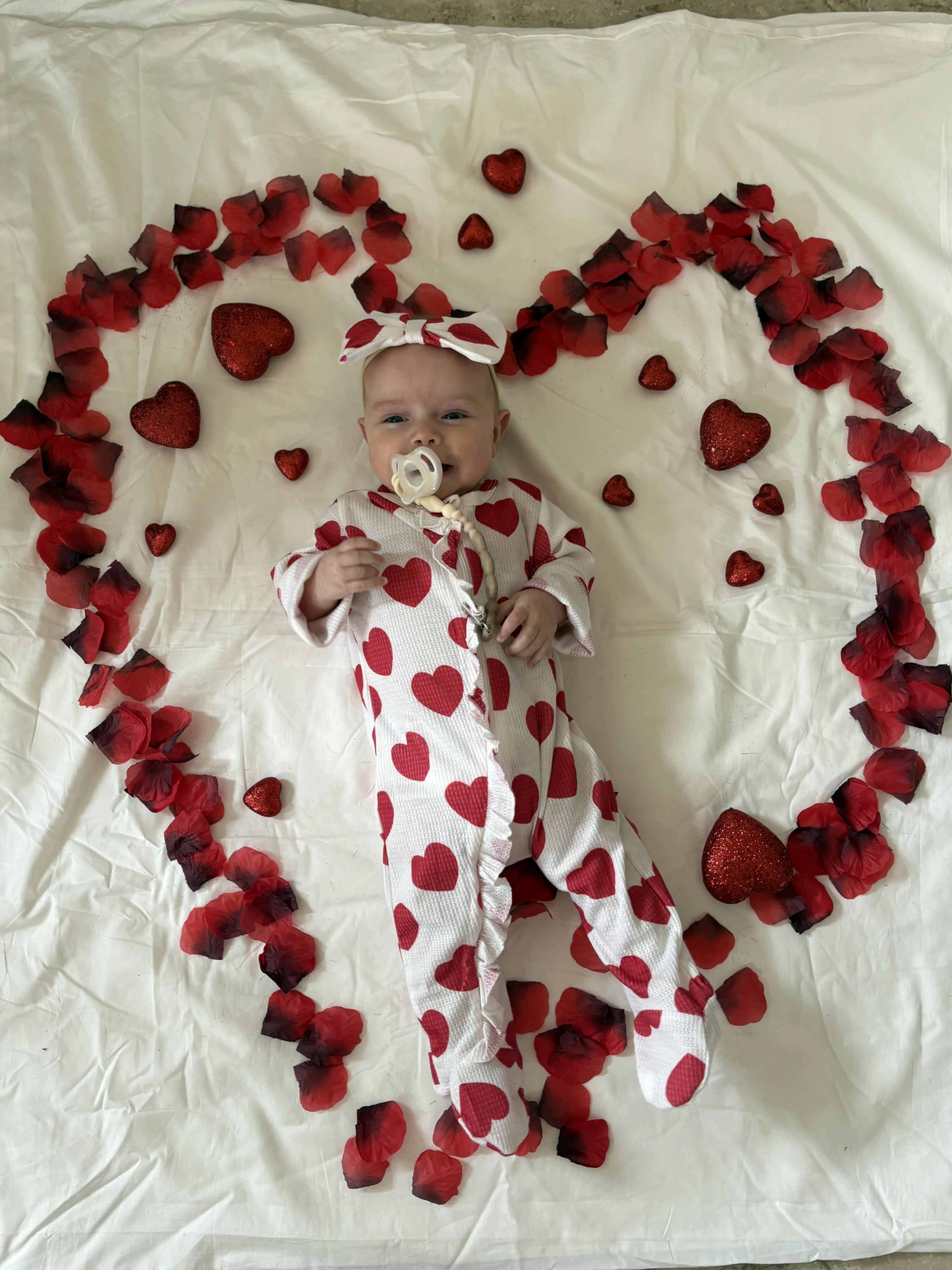 Child in heart-patterned outfit with pacifier, surrounded by red petals and hearts