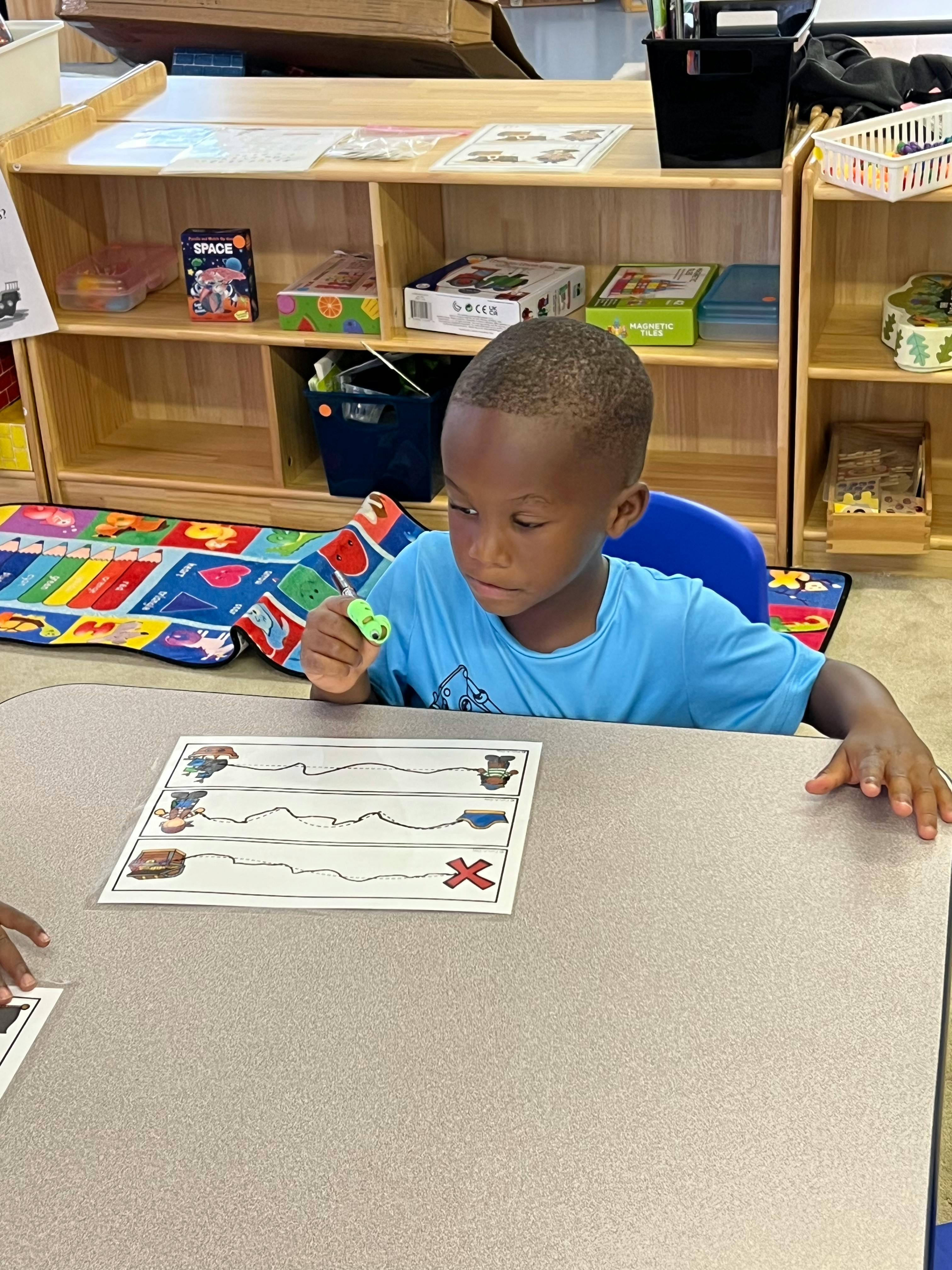 Child in a classroom holding a green object, looking at a worksheet