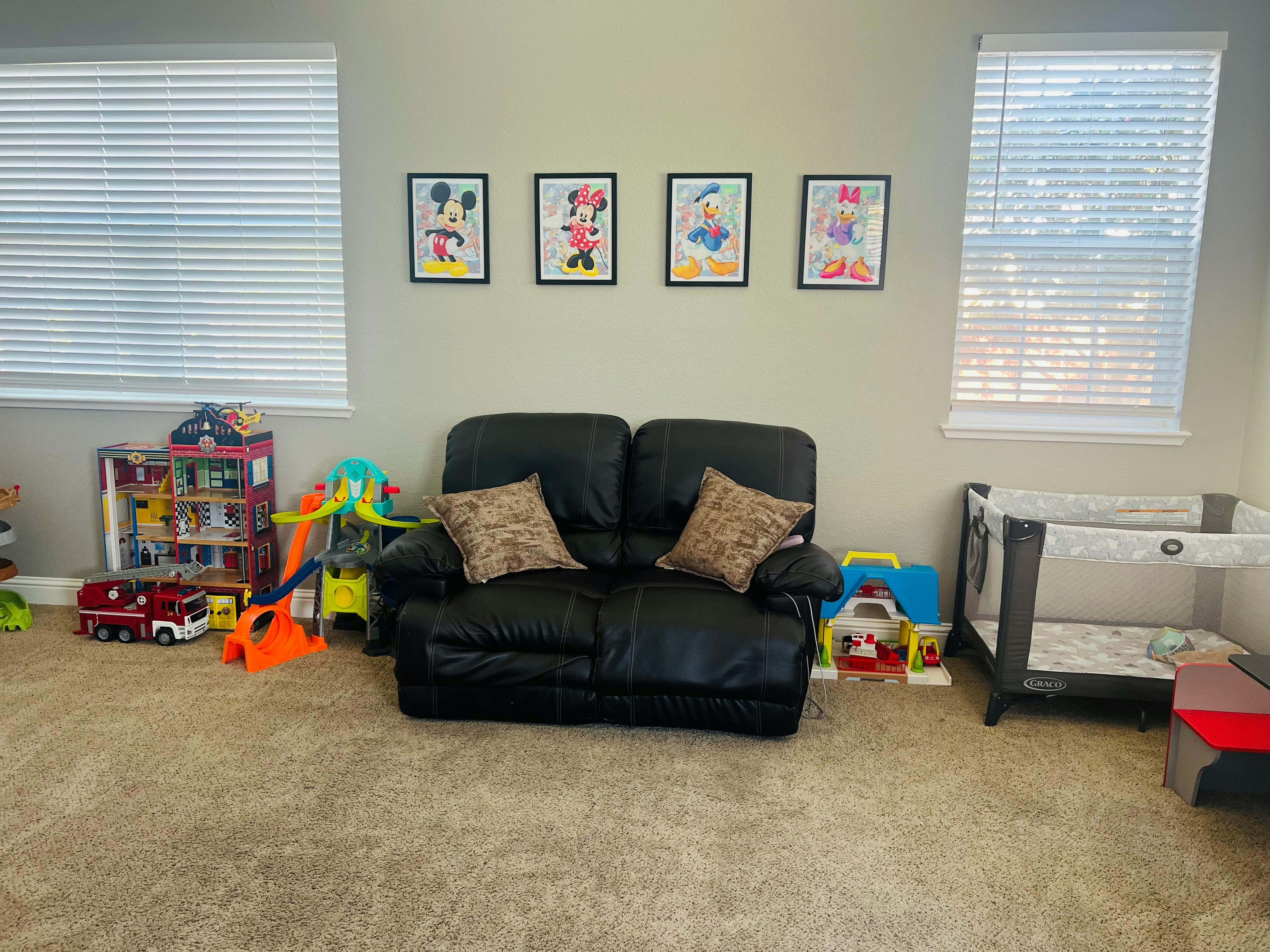 A playroom with a black sofa, a portable crib, and various toys including a fire station