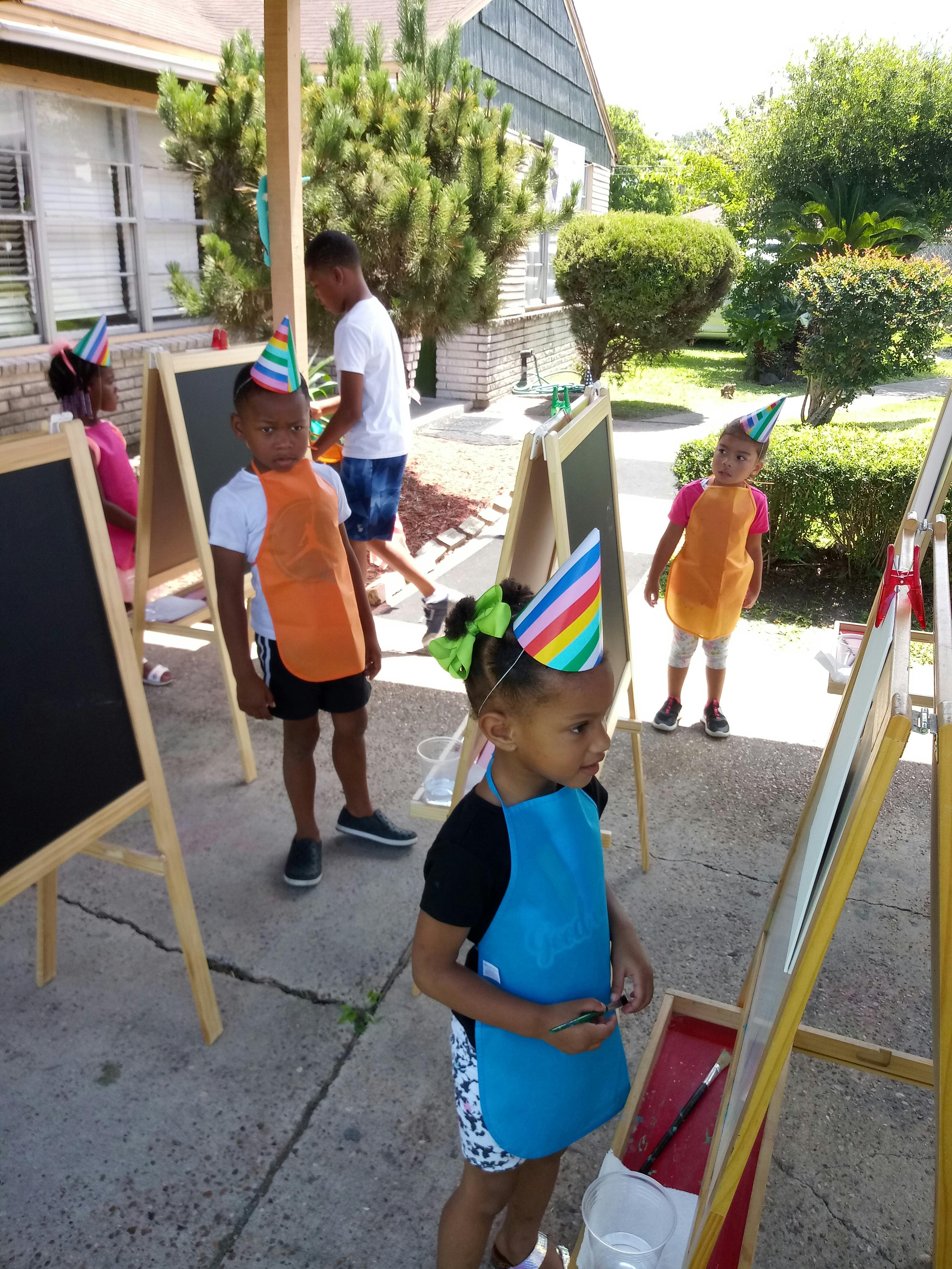 Children wearing party hats and aprons painting at easels outdoors