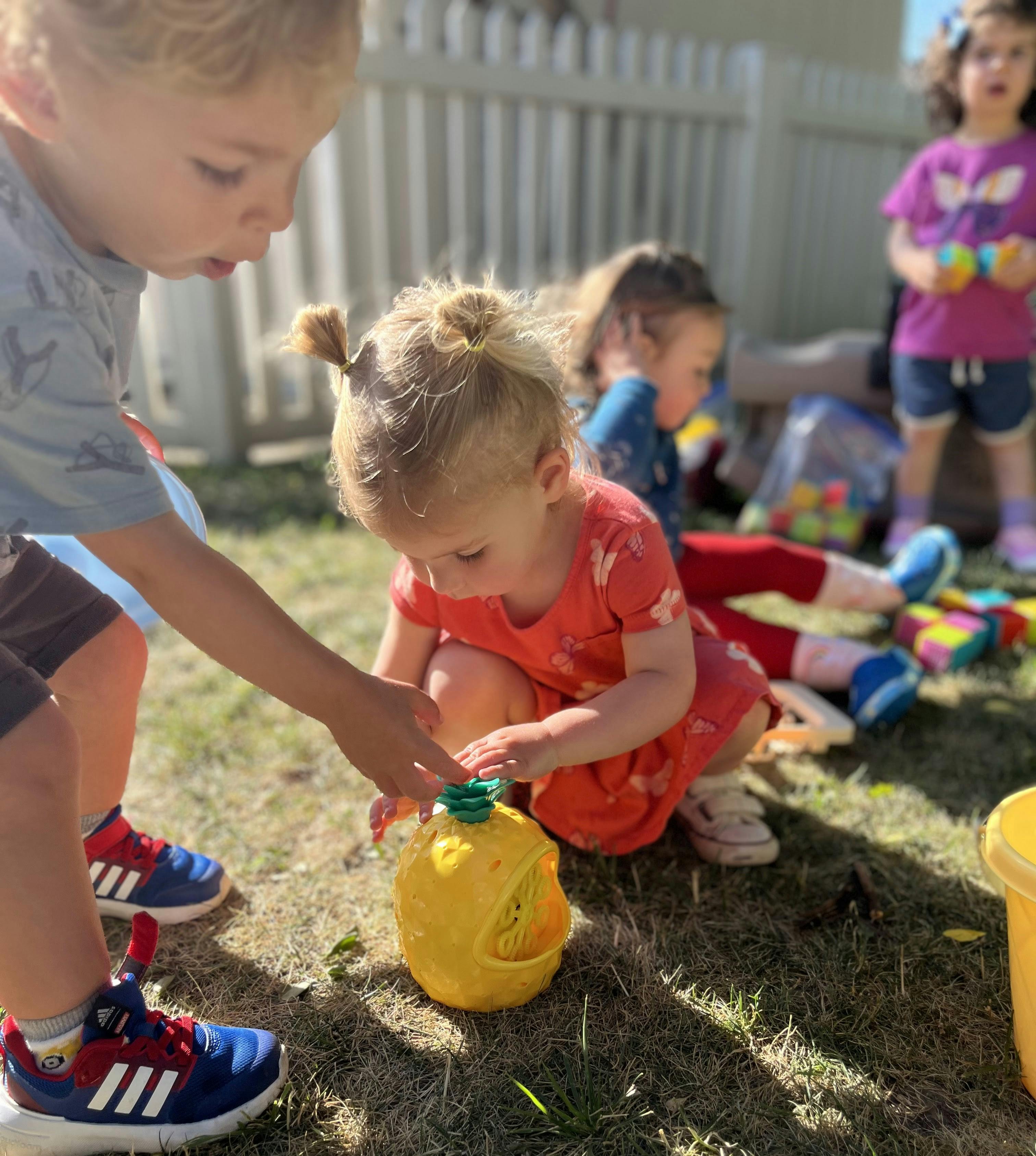 Child playing with a yellow toy on the grass with other children nearby